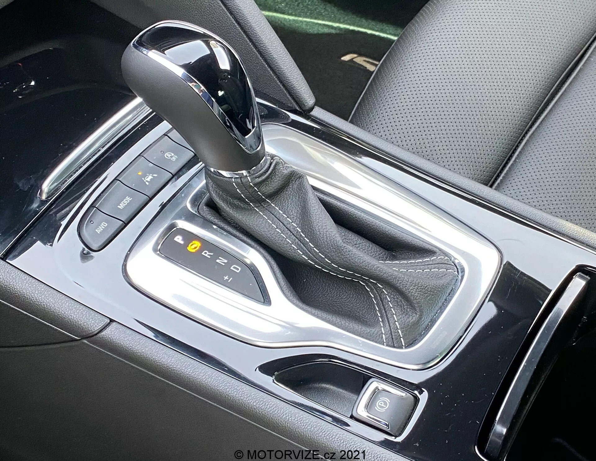 Närbild av den automatiska växelspaken i 2020 Opel Insignia GSi Grand Sport, med den eleganta mittkonsolen sedd ovanifrån. Växelspaken, med sin metalliska finish, är i 'P'-läget och omges av en glansig svart konsol med silveraccenter. Synliga knappar och upplysta indikatorer föreslår moderna elektroniska kontroller och körlägen.