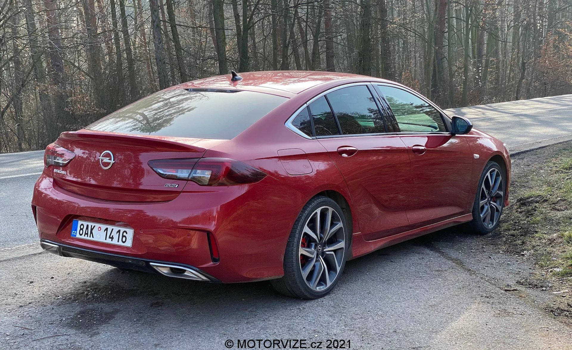 Bilden visar en röd Opel Insignia 2020 (Insignia GSi Grand Sport) sedan parkerad vid vägkanten omgiven av träd, vilket antyder en lantlig eller parkliknande miljö. Fågelperspektivet bakifrån visar bilens eleganta och sportiga design, inklusive den sluttande bakrutan, moderna LED-bakljus, multispektrade legeringsfälgar, dubbla avgasrör, bakre parkeringssensorer och hajfensantenn. Markfrigången är standard för en sedan och balanserar daglig användbarhet med en sportig konfiguration.