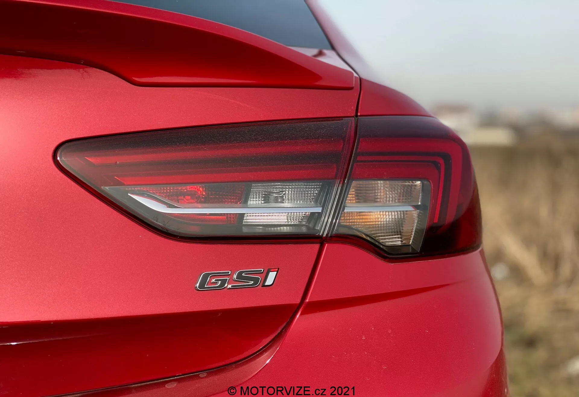 Närbild av bakre vänstra sidan av en Opel Insignia GSi Grand Sport 2020, som visar den moderna designen av den bakre LED-lampan med kombination av bromsljus, blinkers och eventuellt backljus. Den bakre lampan sveper runt hörnet på fordonet, vilket förbättrar synligheten. Under baklyktan är märket 'GSi' tydligt synligt och indikerar modellens utrustningsnivå. Bilen har en glänsande röd lackfinish med aerodynamiska karosslinjer. Fotot fokuserar på bakre lamphuset med ett grunt skärpedjup, vilket framhäver designens detaljer medan de omgivande områdena suddas ut.