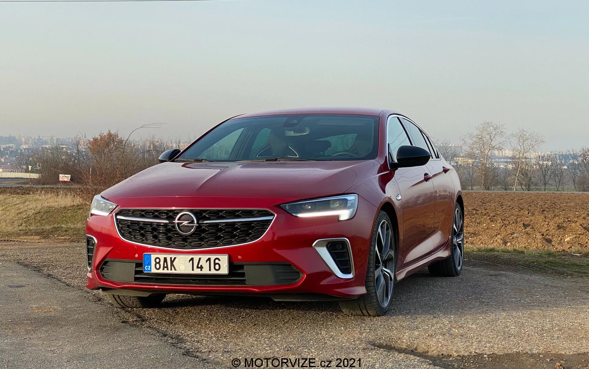Främre vänstra kvartalsvy av en Opel Insignia GSi Grand Sport 2020 från låg vinkel, med sin framträdande grill med logotyp, eleganta LED-strålkastare och dimljus med integrerade dagsljus. Bilden framhäver de skarpa linjerna på huven, frontsensor under registreringsskylten, skulpterade karosslinjer, integrerade blinkers i sidospeglarna och flerspoked fälgar. Bilen är placerad utomhus med klar himmel i bakgrunden, parkerad på en lätt lutning.