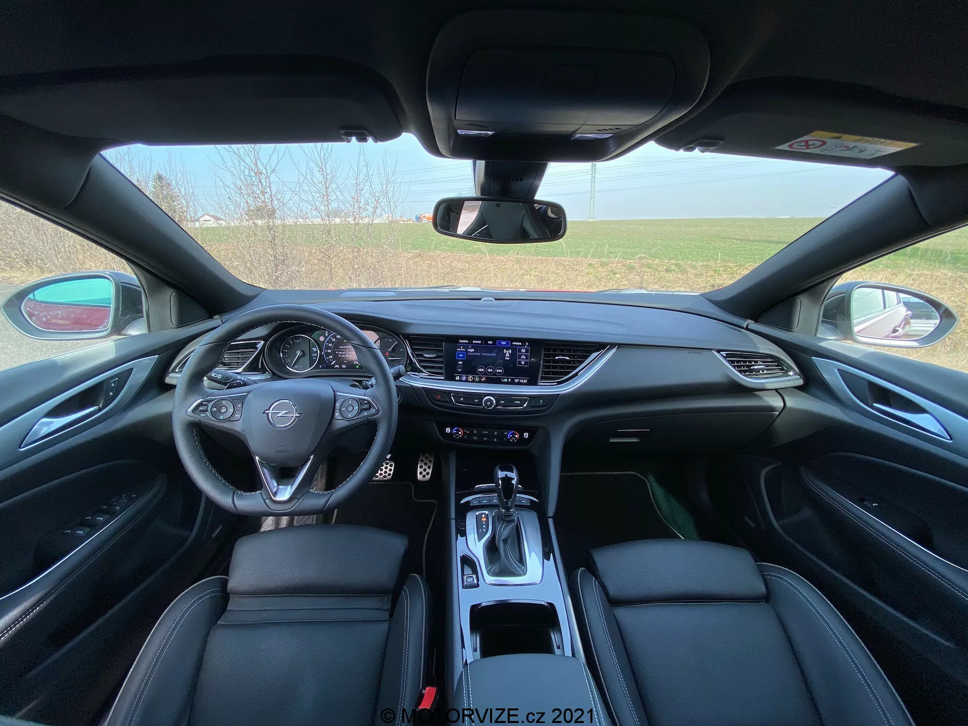 Interiörvy av 2020 Opel Insignia GSI Grand Sport från baksätet perspektiv som visar en detaljerad vy av den främre instrumentbrädan. Bilden är centralt inramad med rattens multifunktionella knappar och märkesemblem i läder, en digital instrumentpanel och en aktiv infotainmentskärm med pekfunktion. Mittkonsolen med smidiga luftventiler, klimatreglageknappar och en modernt designad växelspak framhäver de teknologiska funktionerna. De lyxiga svarta läder framstolarna med detaljerade sömmar bidrar till bilens interiöra elegans.
