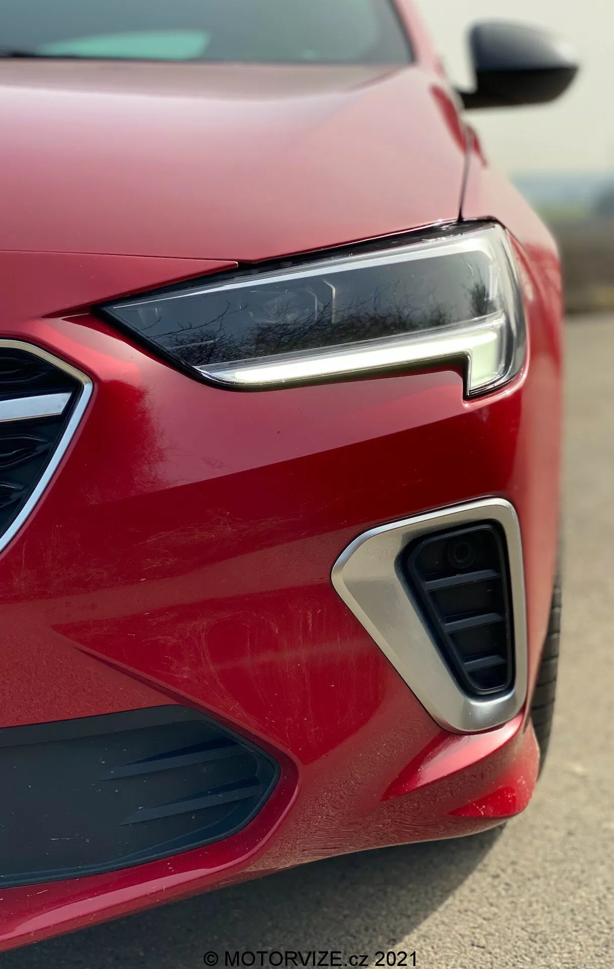 Närbild av det främre hörnet på en modern röd Opel Insignia 2020 (GSI Grand Sport), som visar dess eleganta, smala LED-strålkastare, geometriska grilldesign, luftintag med kromaccent och potentiellt dimljus från en låg vinkel.