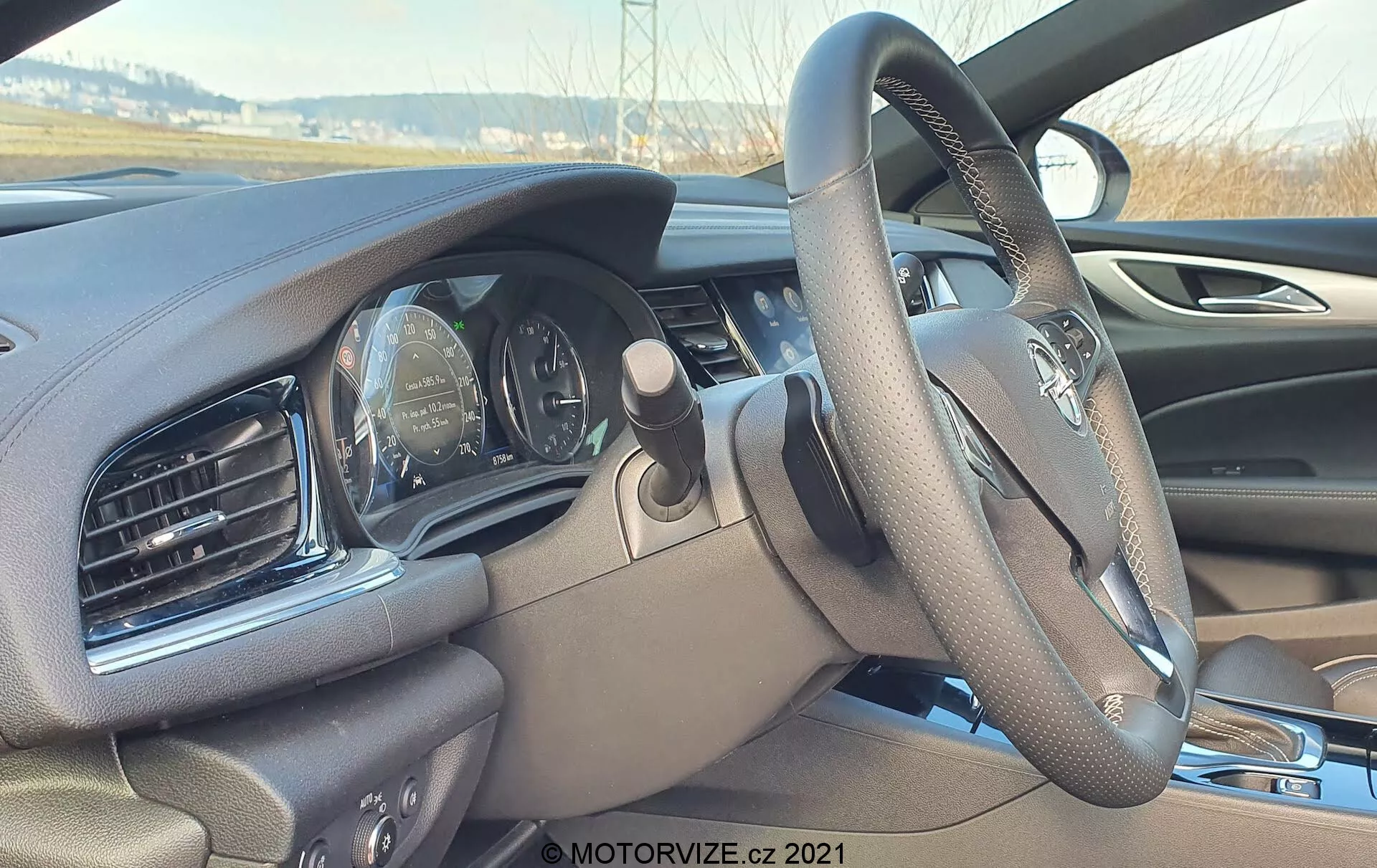 Interiör av Opel Insignia 2020 (Insignia-GSI-Grand-Sport) från passagerarsätet, fokuserar på ratt och instrumentpanel. Vinkeln är något neråtlutande från höger sida, visar en multifunktionell ratt med suddig logotyp och delvis synlig analog instrumentpanel med hastighetsmätare och varvräknare. Panelen har ventilationsöppningar och skulpturerad design, med mörkt material. En skymt av infotainmentskärmen på höger kant.