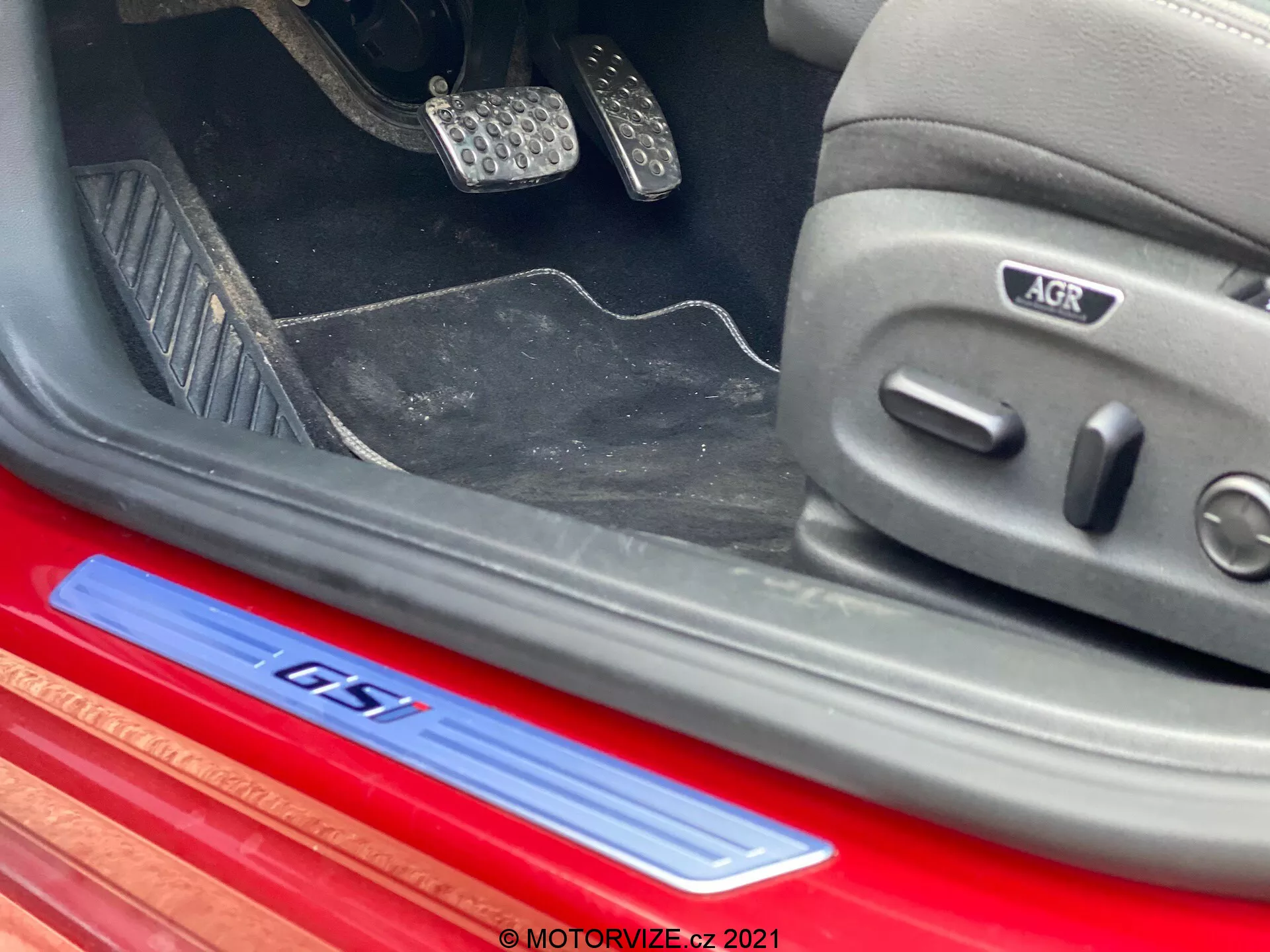 Närbild i vinkel av den nedre delen av interiören i en Opel Insignia GSI Grand Sport 2020. Visade detaljer inkluderar en röd GTI-skylt med silver-och-blå logotyp på dörrtröskeln i borstad aluminium, metalliska sportpedaler med gummiinsatser, delvis synliga mattor och en sidomonterad elektriskt justerbar bilstol med ikoner och AGR-ergonomisk certifieringsmärke. Interiören visar en sportig och premium design med mörka toner och accenter i rött och metall.