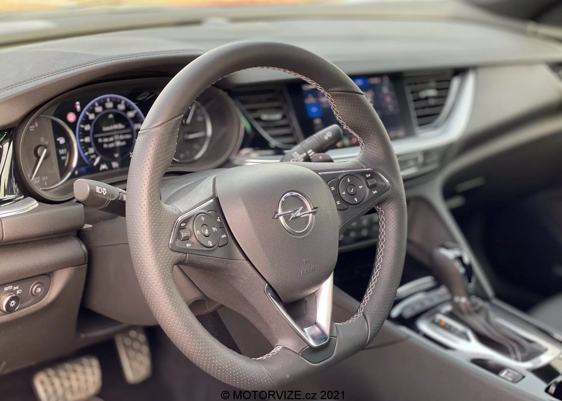 Interiör av Opel Insignia GSi Grand Sport 2020 från förarens perspektiv, med fokus på mittmonterad ratt med kontrollknappar för multimedia och farthållare. Instrumentpanelen bakom ratten inkluderar en hastighetsmätare, varvräknare och en digital display. Delvis synlig mittkonsol med luftkonditioneringsventiler, en infotainmentskärm och klimatanläggningsknappar. Den läderklädda ratten och den moderna designen betonar komfort och teknik.