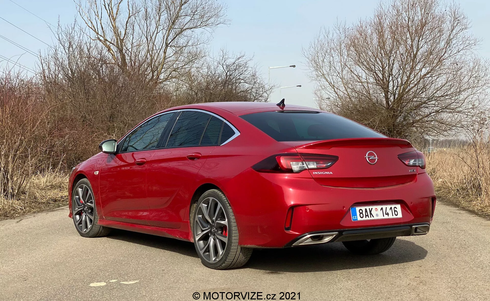 Bilden visar en röd Opel Insignia GSi Grand Sport 2020 från en trekvartsvinkel bakifrån, med betoning på högersidan och bakdelen av fordonet som är placerat diagonalt. Fotot taget i dagsljus på en torr, gräsbevuxen plats med nakna träd, troligen sena hösten eller vintern. Den moderna, eleganta och sportiga sedanen har skarpa karosslinjer, sluttande bakruta, kort baklucka, framträdande bakspoiler, stora flerradiga aluminiumfälgar, dubbla avgasrör och en märkbar bakre diffusor. De bakre LED-ljusen, det centrala logotypen på bakluckan, modellens emblem och den centralt placerade nummerskylten är synliga. De tonade fönstren och sidospeglar i karossens färg bidrar till bilens aggressiva men eleganta hållning.