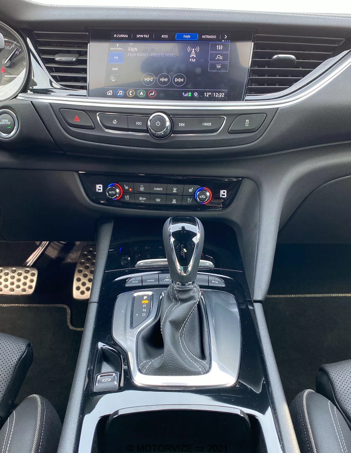 Bild på interiören av 2020 Opel Insignia GSi Grand Sport som visar främre kabinområdet från förarens eller passagerarens perspektiv. Fotot fångar mittkonsolen från växelspaken upp till infotainmentskärmen. Växelspaken är i parkeringsläge, vilket tyder på en automatisk växellåda, och är omgiven av en glansig svart kant. Nära finns det knappar för körlägen och en lädermanschett runt växelspaken som bidrar till en premiumkänsla. Bakom växelspaken finns två mugghållare och ett förvaringsutrymme med ett utdragbart lock. Ovanför sitter klimatkontroller med dubbelzonstemperaturindikatorer och fysiska knappar. Längre upp finns det aktiverade infotainmentsystemet som visar en navigationskarta och andra inställningar. Instrumentbrädan är av mjuk plast och integrerar ventiler på vardera sidan om infotainmentskärmen sömlöst.