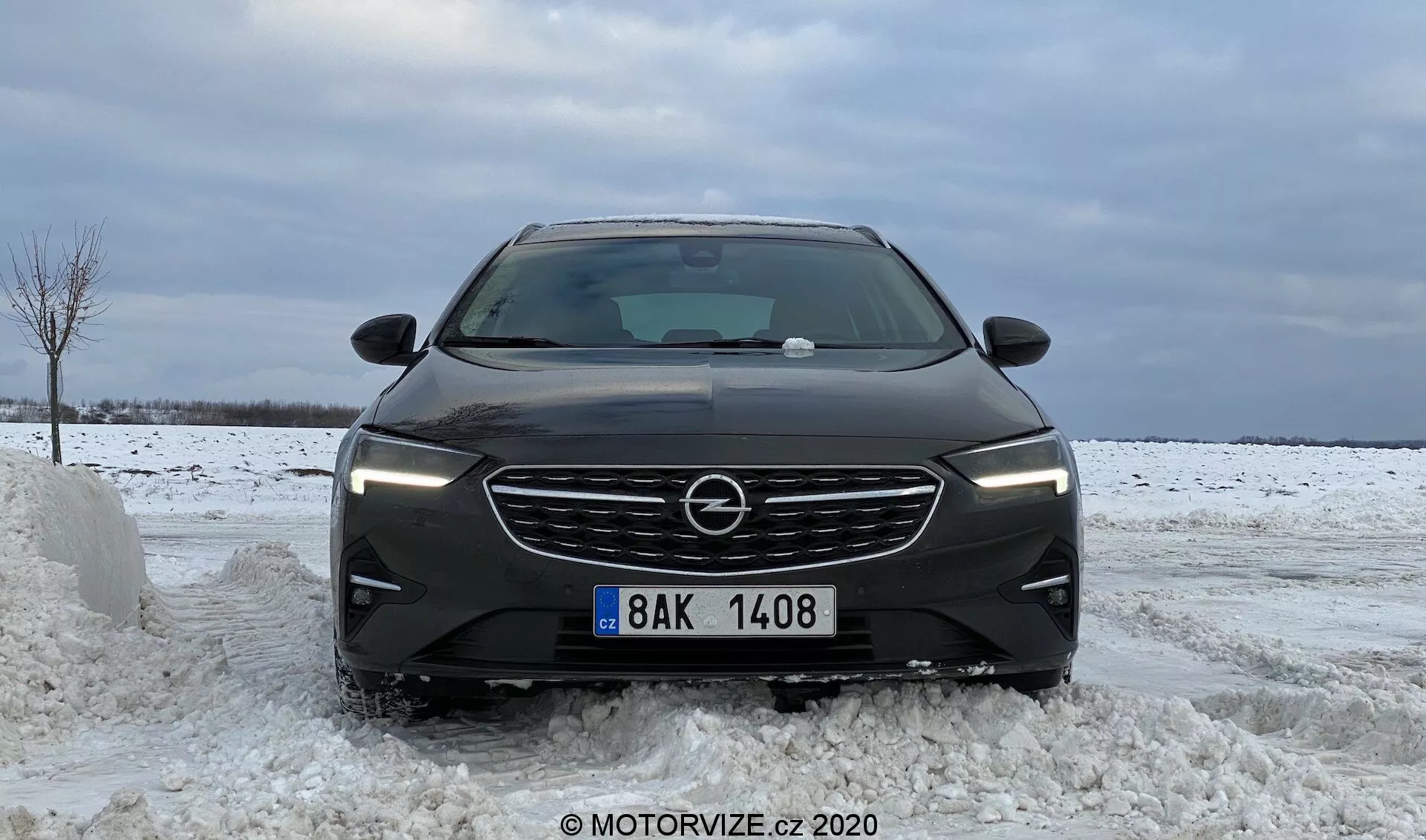 Frontal vy av en 2020 Opel Insignia Sports Tourer på en snöig väg som visar dess moderna designinslag såsom horisontella ribbor på grillen, vinklade LED-strålkastare, skulpterade konturer på motorhuven, bred främre stötfångare med integrerade luftintag och centrerad registreringsskylt, som betonar vinterförhållanden.