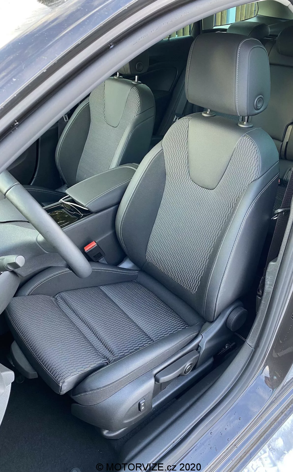 Närbild, vertikal bild av interiören i Opel Insignia 2020 (Insignia Sports Tourer) fokuserad på förarsätet med en blandad design av tyg och syntetiskt läder, manuella sätesjusteringskontroller, delvis synlig multifunktionell ratt och höjdjusterbara säkerhetsbälten, som framhäver ergonomiska och klädsel relaterade egenskaper.