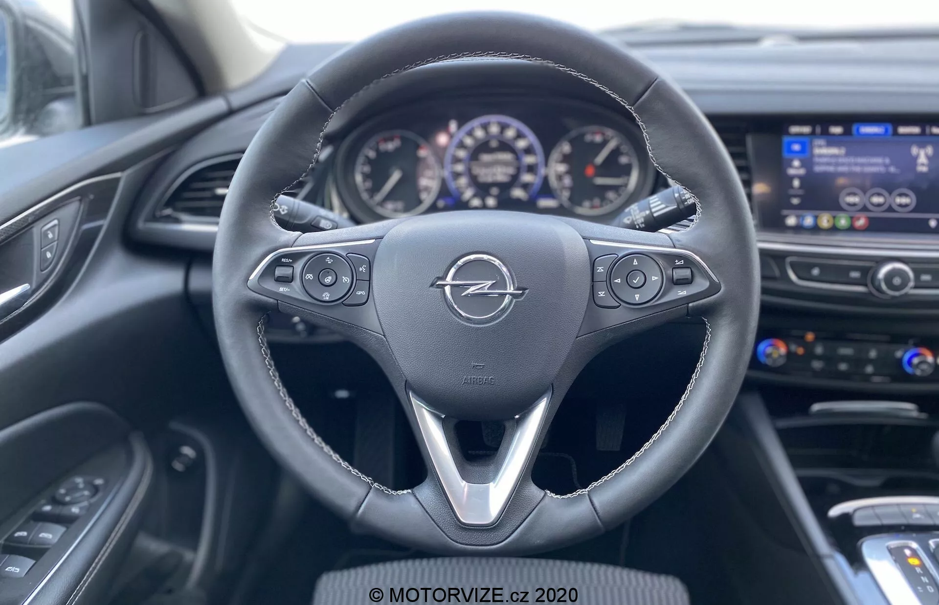 Utsikt från förarsäten i 2020 års Opel Insignia Sports Tourer som visar den läderklädda multifunktionsratten med tre-ekers design, infotainmentsystemets kontroller, farthållarinställningar, delvis synlig instrumentpanel med analoga urtavlor och liten digital display, infotainmentdisplayen på mittkonsolen, luftventiler, klimatanläggning med knappar och vred, mörkfärgad instrumentpanel med strukturerat mönster samt delvis synlig förardörrs kontrollpanel.