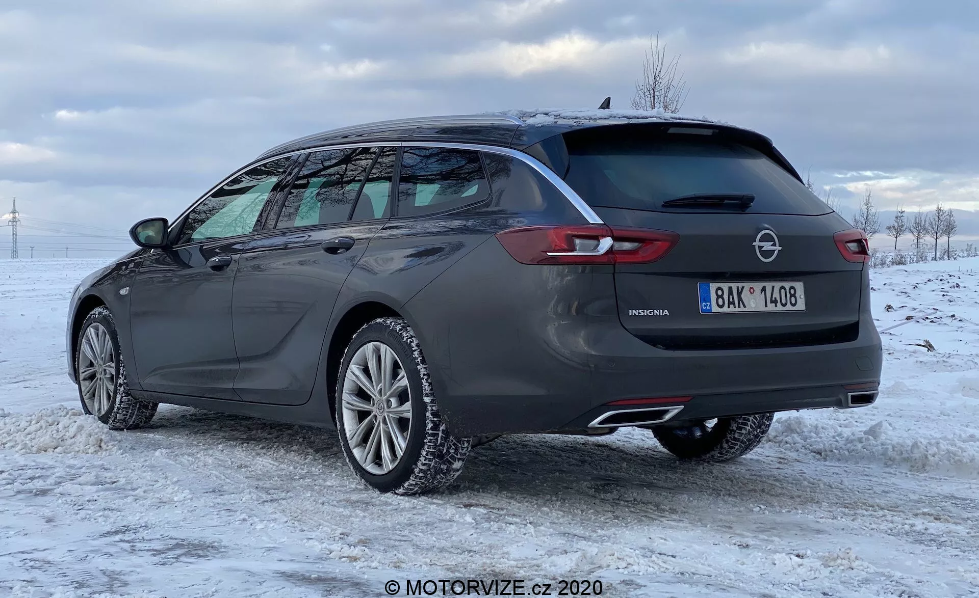 Sido- och bakvy av en mörkfärgad 2020 Opel Insignia Sports Tourer i ett snötäckt landskap, som visar dess moderna, eleganta design. Bilen har lättmetallfälgar, takräcken, dubbla avgasrör, LED-bakljus, en bakspoiler, en bakre vindrutetorkare och en hajfennaantenn. Den här bilden framhäver den stora bagagekapaciteten, sportiga estetiken och praktiska funktioner hos den moderna modellen.