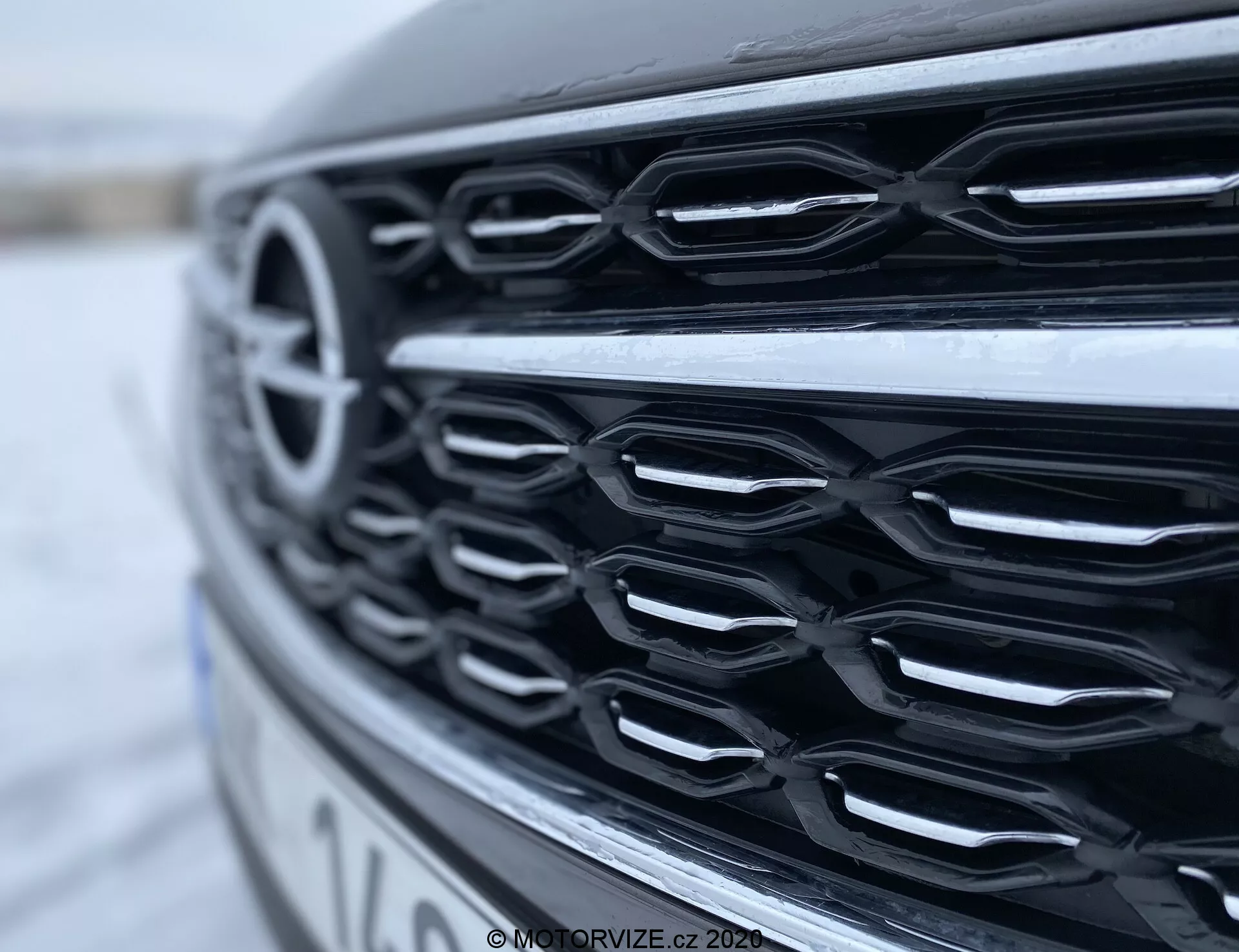 Närbild från sidan av främre grillen på Opel Insignia Sports Tourer 2020 som visar kromliknande horisontella ribbor med en lätt böjning, fotograferad från en låg vinkel. Reflektionerna på den blanka finishen syns i diffust naturligt ljus och framhäver grillens konturer och djup, medan den suddiga bakgrunden betonar dess moderna design.