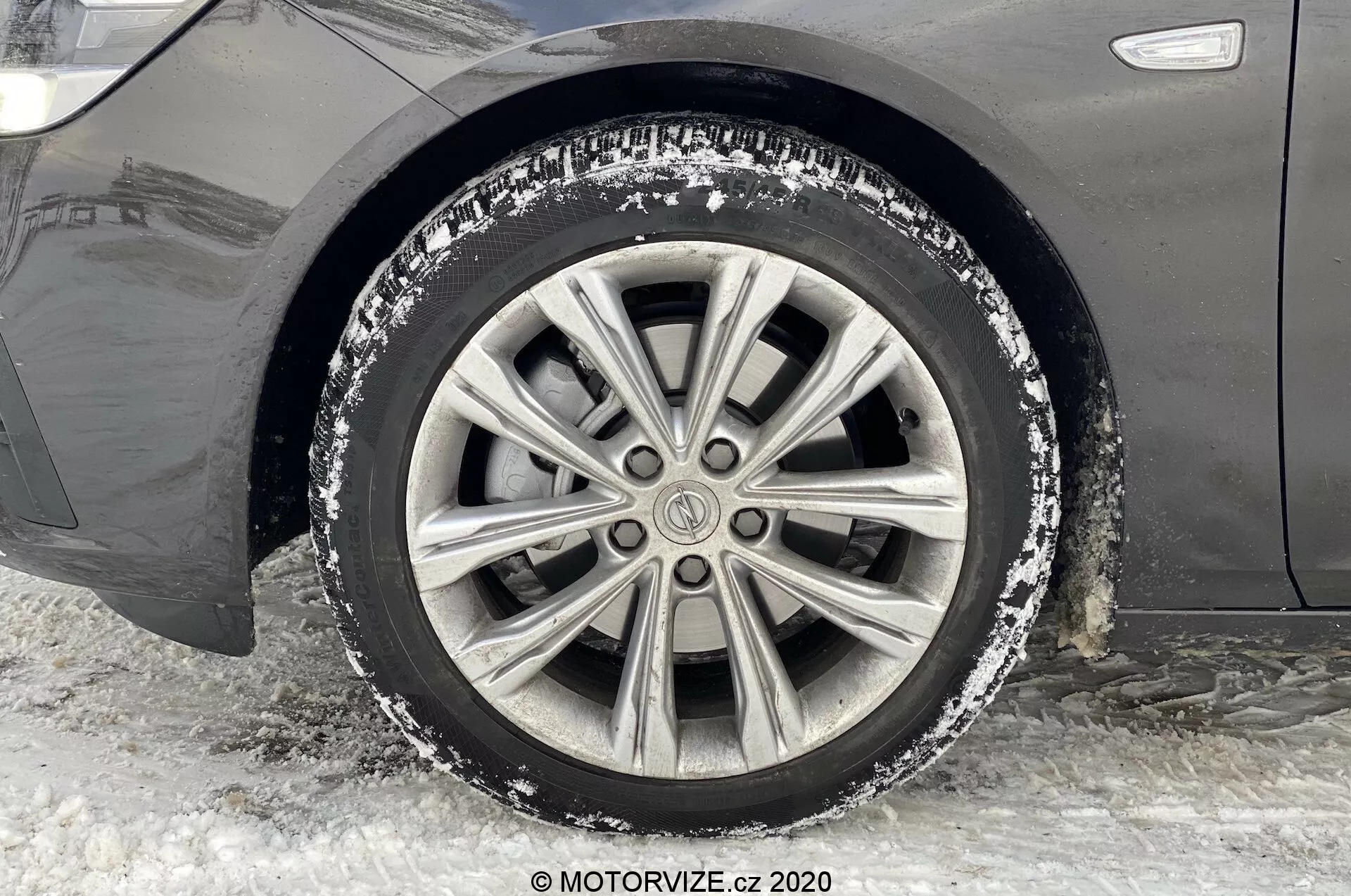Närbild av framhjulet och skärmen på passagerarsidan av en Opel Insignia 2020 (Insignia Sports Tourer). Bilden är tagen i marknivå och fångar hjulets sidoperspektiv. Bilen är parkerad på komprimerad snö och is, vilket tyder på en vintermiljö. Hjulet har en flerskiktslegerad fälg med lite snö och vägsalt, vilket visar på nyligen körning på vintervägar. Mindre repor och smuts är synliga på fälgen, vilket speglar användning i snöiga förhållanden.