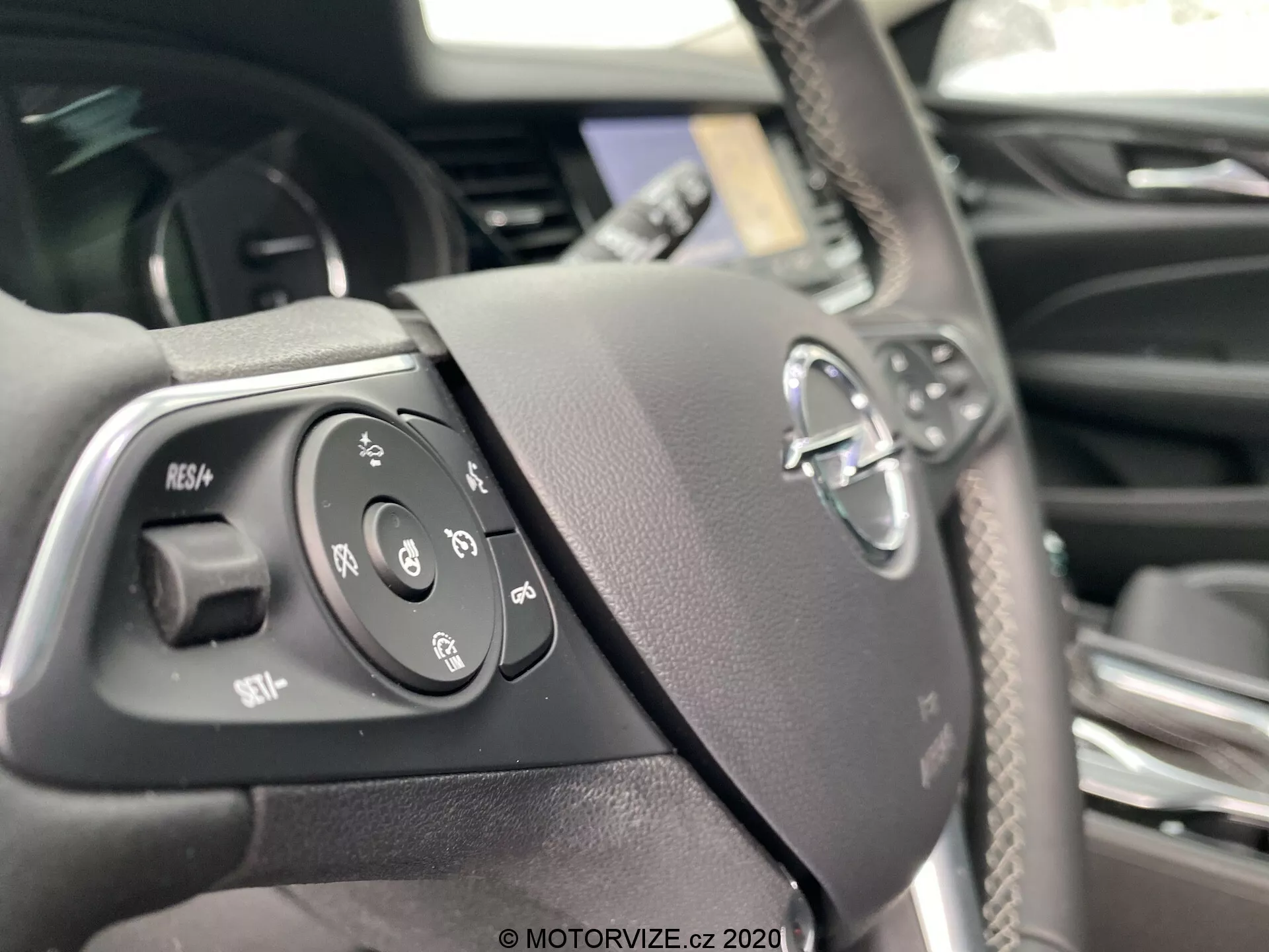 Närbild av ratten i en Opel Insignia Sports Tourer 2020 från förarens perspektiv som visar integrerade kontroller till vänster, inklusive farthållare, röstkommando, telefonfunktioner och ljudkontroller. Ratten har taktila knappar för enkel manövrering och ett suddigt tillverkningsmärke i mitten, placerat i en premiuminteriör med kolfiberliknande trim.