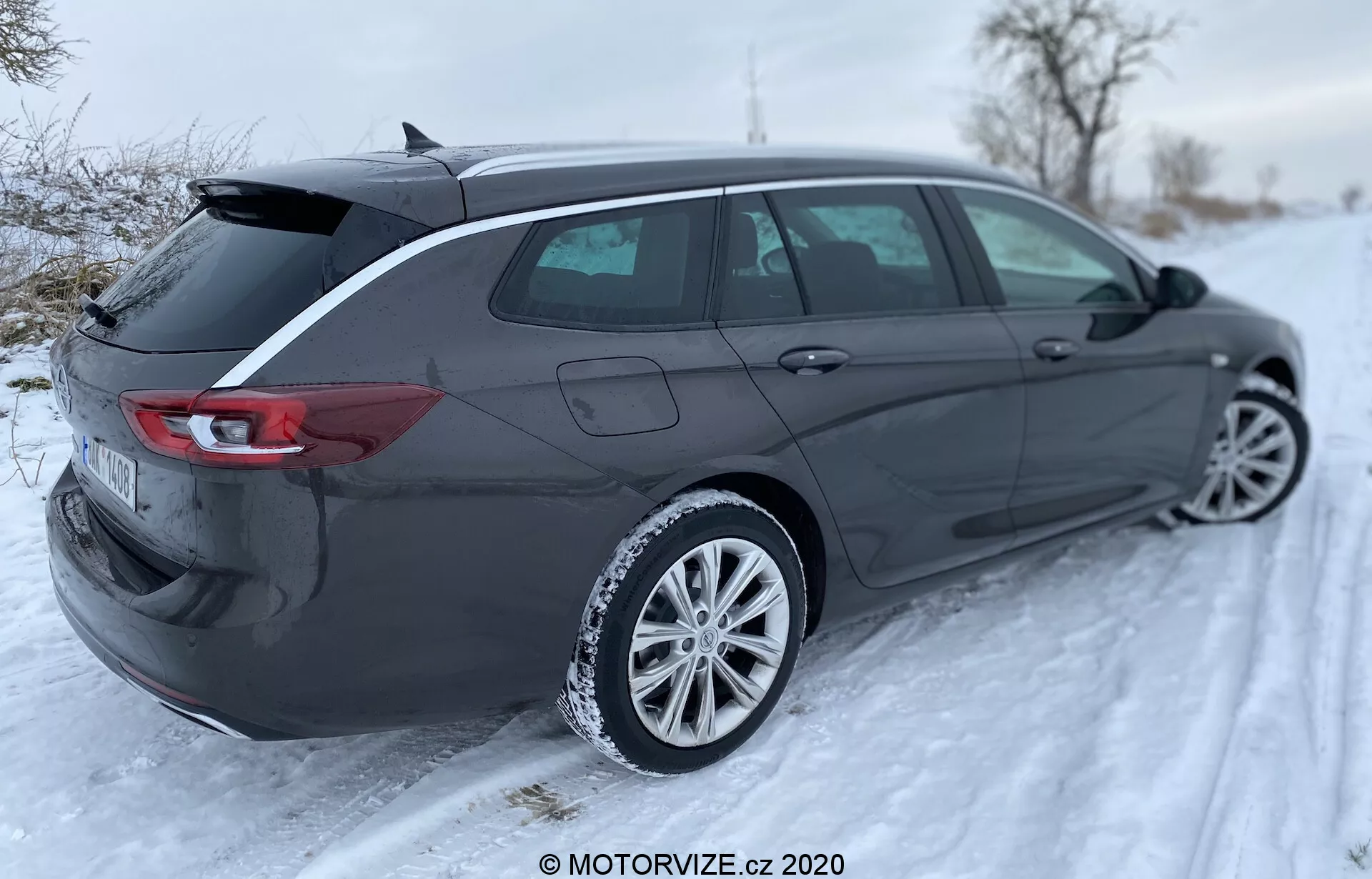 Opel Insignia Sports Tourer 2020, en elegant och modern kombi, är avbildad i en vintermiljö utomhus från ett trekvart bortifrån, vilket framhäver dess aerodynamiska profil, baklucka, baklyktor, bakre stötfångare, sidospeglar, lättmetallfälgar och distinkta karosslinjer. Den öppna bagageluckan indikerar gott om lastutrymme och det snöiga landskapet visar bilens potential i vinterförhållanden. Baklyktorna har en horisontell LED-design som sveper runt sidorna, registreringsskylten är suddig och bilden har en MOTORVIZE.cz 2020 vattenstämpel.