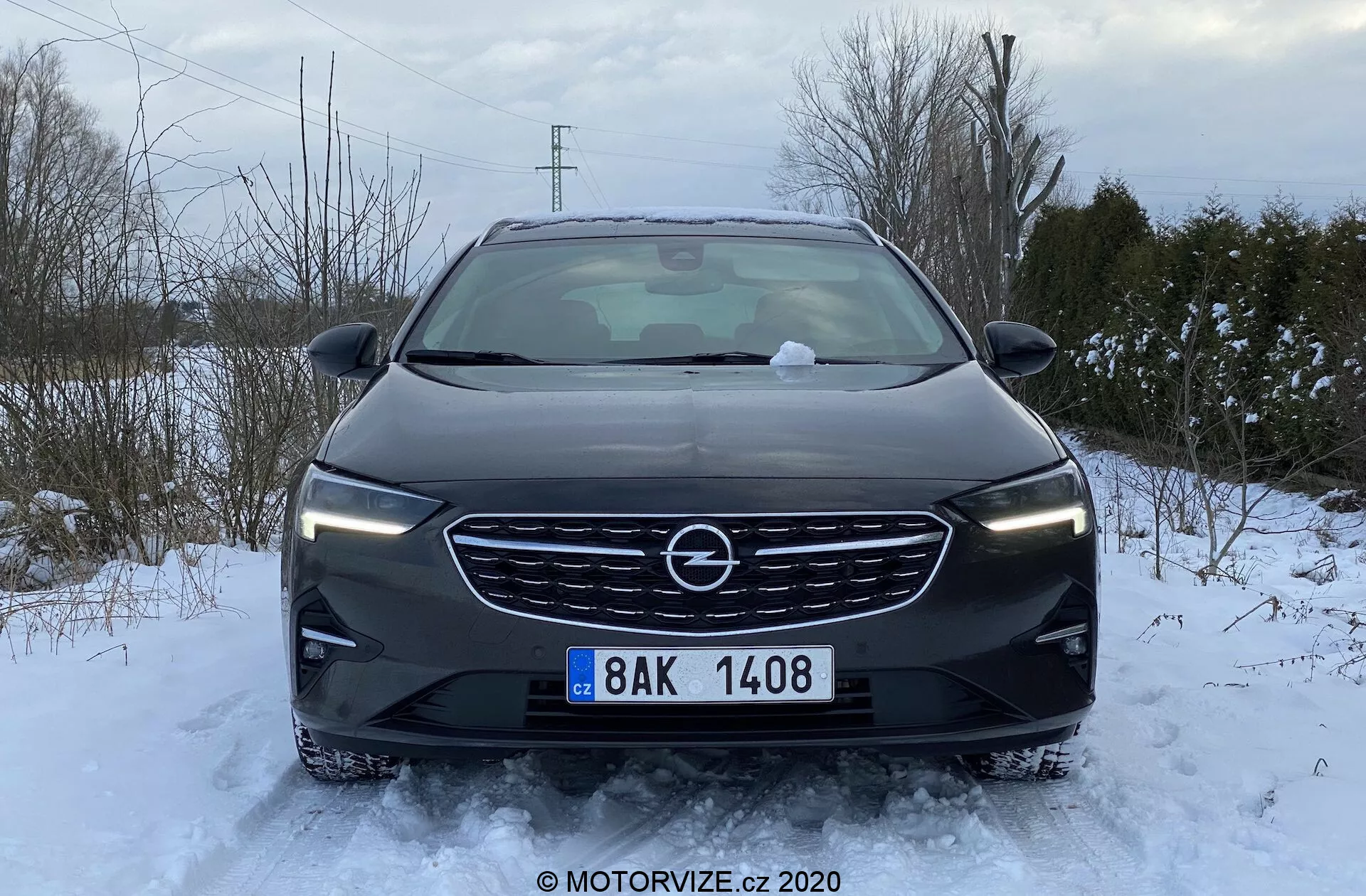 Frontal vy av en stillastående 2020 Opel Insignia Sports Tourer i en snöig miljö. Motorhuv, grill, strålkastare och främre stötfångare är synliga, med snö på marken och bilen. Grillen har ett framträdande centralt märke och en horisontell lameldesign. Snyggt utformade strålkastare sträcker sig horisontellt, medan stötfångaren inkluderar dimljus, luftintag och parkeringssensorer. Sidospeglarna är utfällda, vilket indikerar att bilen är klar för körning. Molnigt dagsljus ger en jämn belysning utan skarpa skuggor.