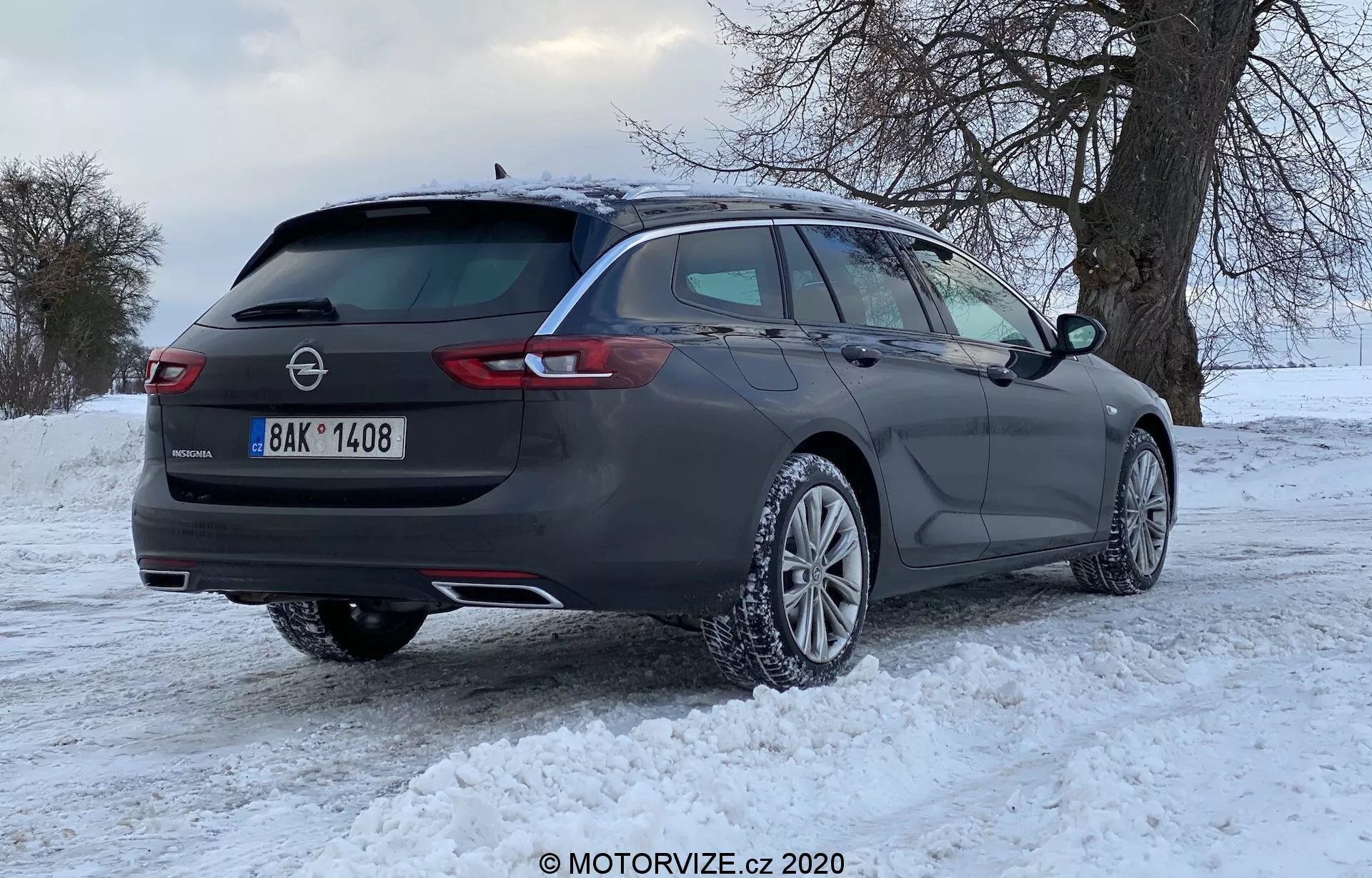 Mörkgrå Opel Insignia 2020 kombi med fler-ekrade lättmetallfälgar på snötäckt mark, bakre trekvarts vy som visar horisontella LED-bakljus, takräcken, tonade fönster och märken som anger märke och modell, med vintrig bakgrund av lövfria träd och molnig himmel.
