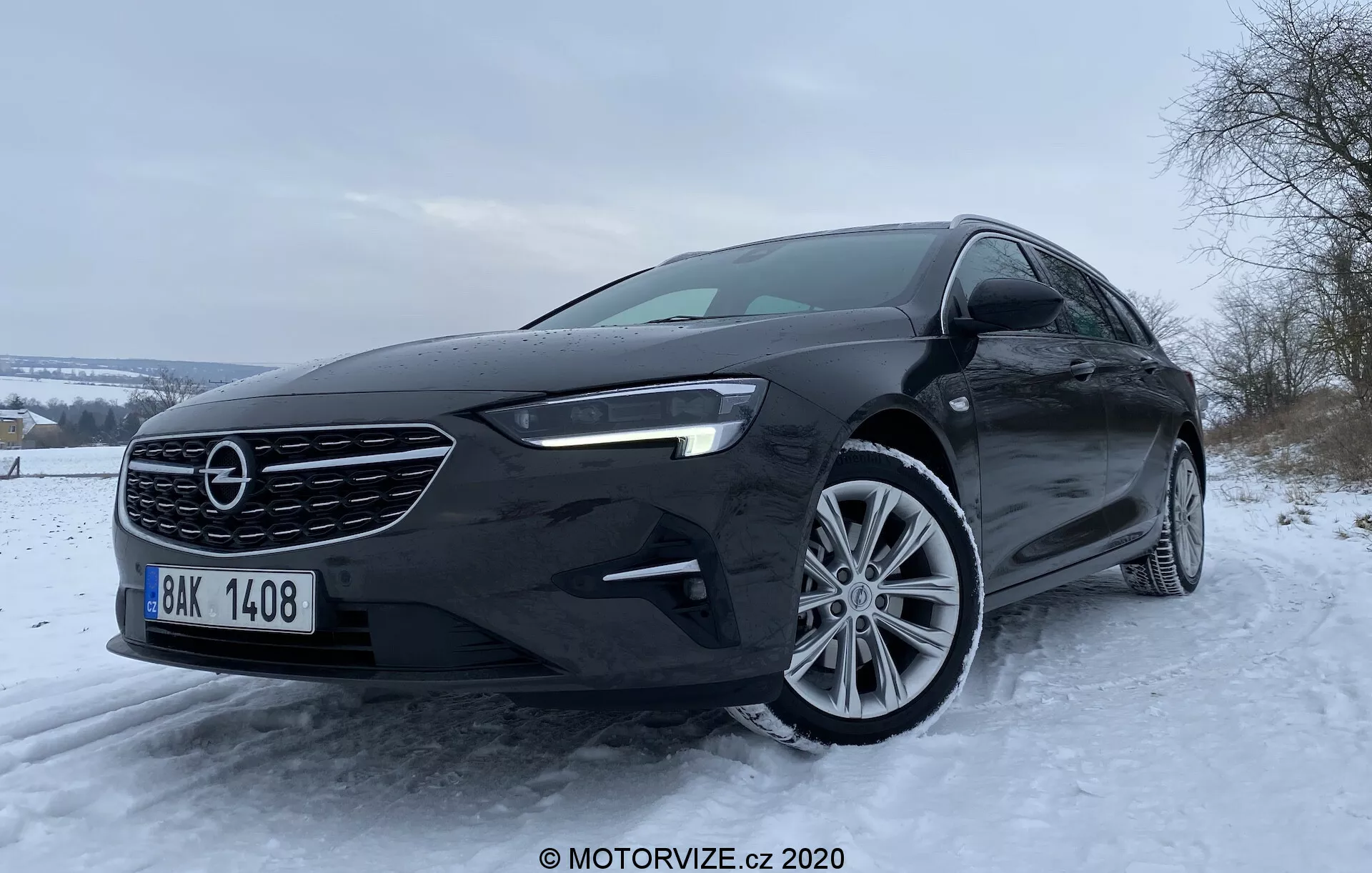 Bilden visar en 2020 Opel Insignia Sports Tourer sedan från en låg, frontvinkel som framhäver dess distinkta grill, främre stötfångare, LED-dagsljus och lättmetallfälgar, mot en vintrig bakgrund med snötäckta träd och molnig himmel.