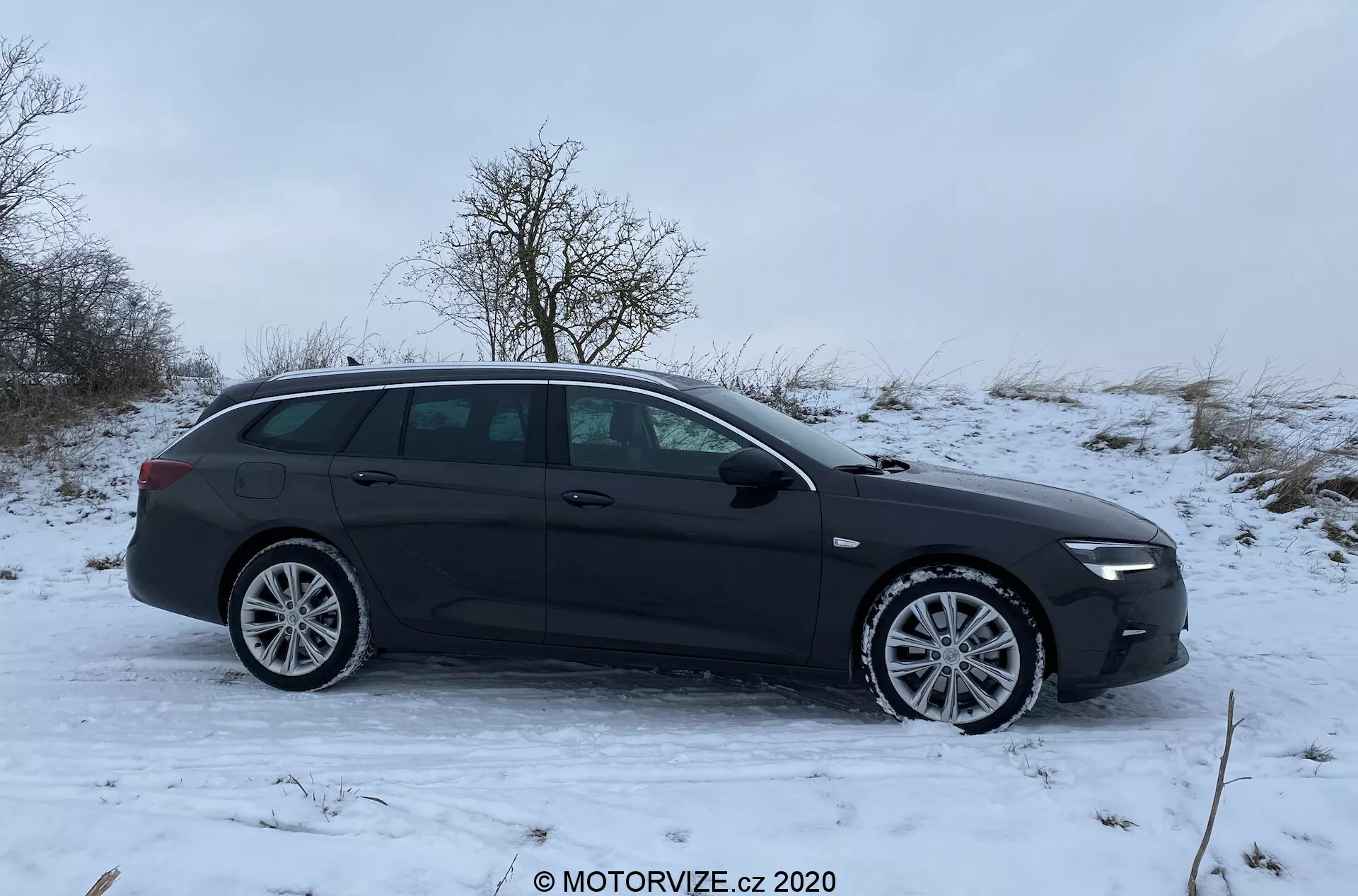 Bild av en svart Opel Insignia 2020 kombi parkerad i en lätt lutning med snötäckt mark, sedd från sidoprofilvinkel. Hela vänstra sidan av fordonet är synlig, inklusive flerspåriga lättmetallfälgar, fyra dörrhandtag, sidobackspeglar, fönster och takräcken. Bilens moderna design har släta konturer och en elegant taklinje, vilket antyder användbarhet för att bära föremål som takräcken eller sportutrustning.