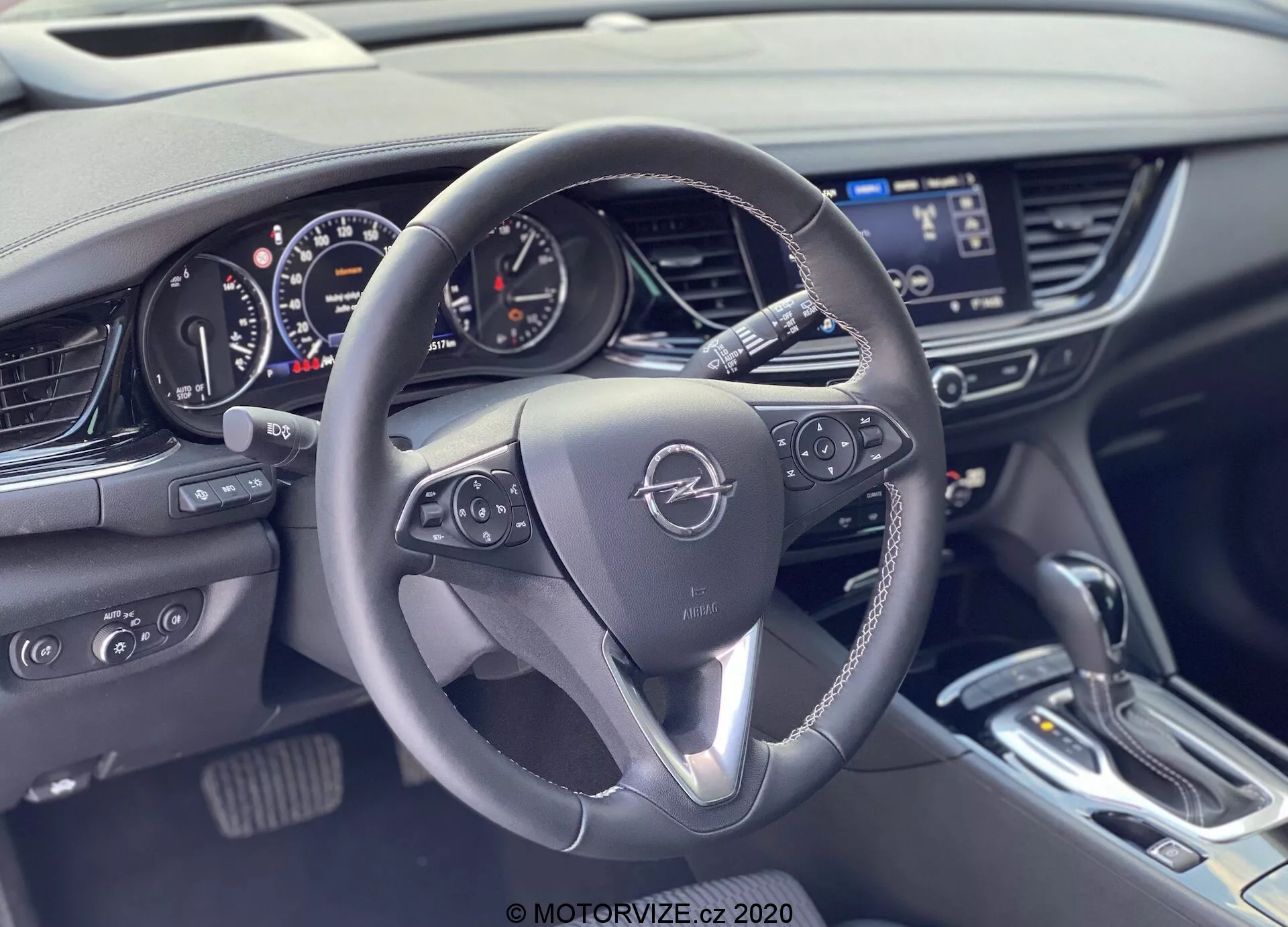 Interiörvy av en 2020 Opel Insignia Sports Tourer från baksätena, något förskjuten mot förarsidan, med fokus på ratten och instrumentbrädan. Den treekrade ratten har metalliska accenter och kontrollknappar för infotainment och fordonsinformation. Emblemet är framträdande i mitten. Bakom ratten finns en kombination av analoga och digitala instrument med en hastighetsmätare, varvtalsmätare och digitala skärmar. Till höger finns mittkonsolen med en infotainmentskärm, klimatkontroller och en elegant växelspak. Interiören inkluderar läder, metalliska accenter och mjuka material, vilket visar en modern, teknikorienterad design.