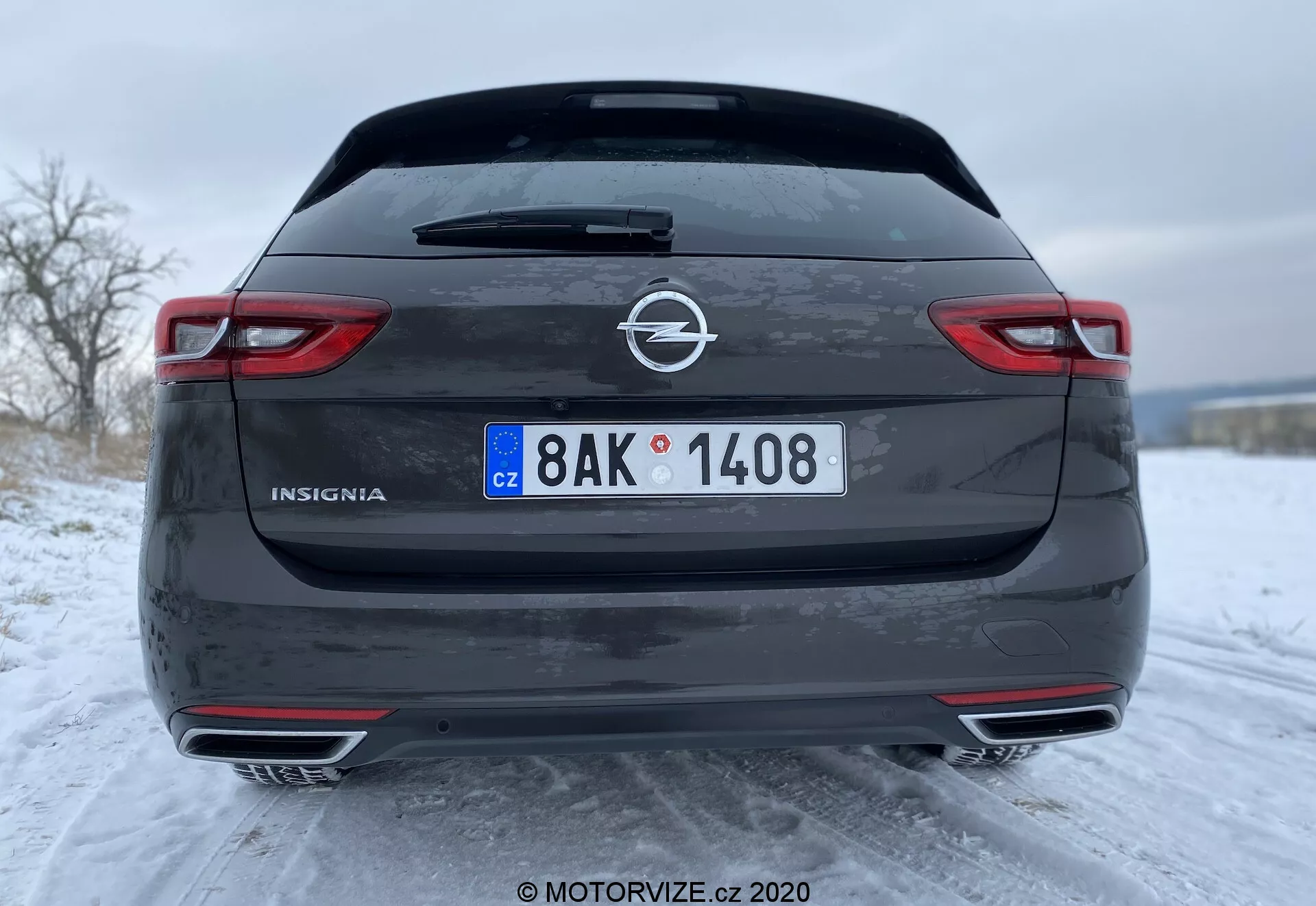 Bakifrånvy av 2020 Opel Insignia Sports Tourer i mörkgrå eller svart, som visar den detaljerade designen med långsträckta horisontella bakljus, dubbla avgasrör och synliga parkeringssensorer på den nedre stötfångaren. Fotot lyfter fram fordonets sportiga egenskaper, den europeiska registreringsskylten och den snöiga vintermiljön.