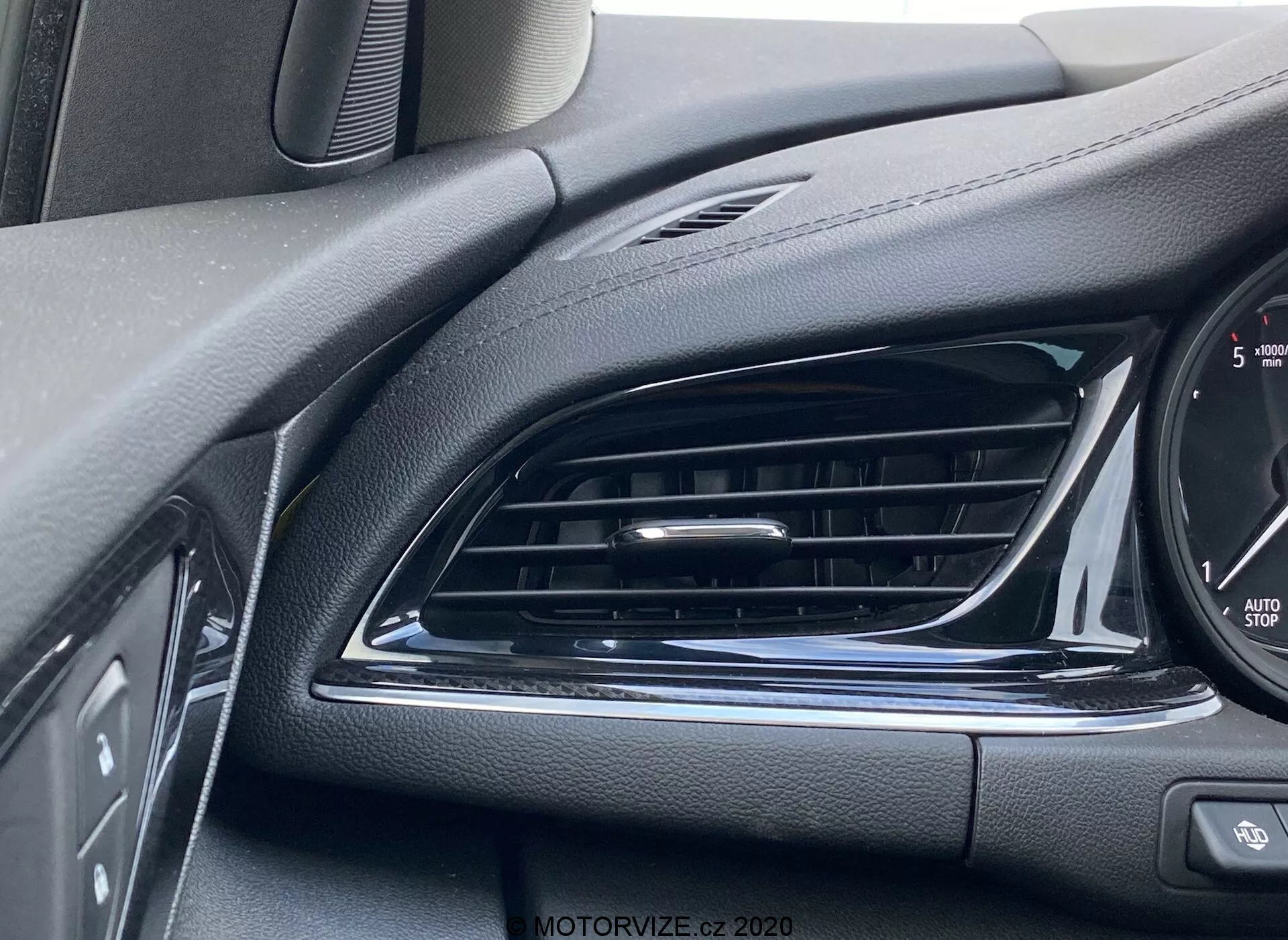 Närbild av interiören på Opel Insignia Sports Tourer 2020. Innehåller en svart luftventil med horisontella lameller, strukturerade ytor som liknar läder för estetik och bländskydd, glänsande kromaccent, delvis synlig kontrollpanel med knappar, kanten av instrumentklustret med hastighetsmätartal och texten 'Auto Stop' som indikerar automatiskt start-stoppsystem. Vinklat skott från förarens sida som fokuserar på luftventilen och omgivande detaljer.