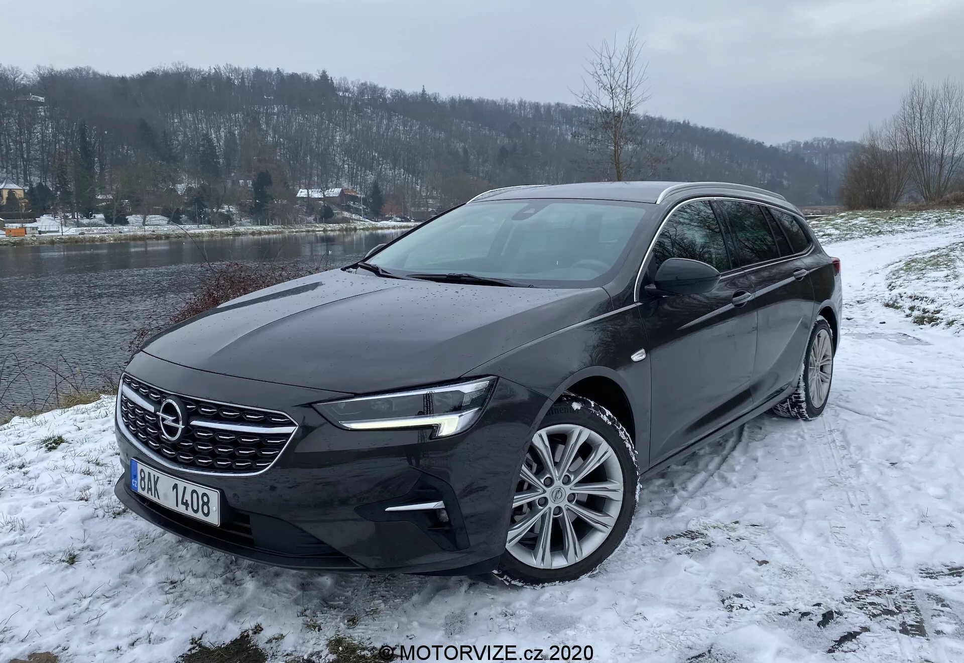 2020 Opel Insignia Sports Tourer i en vintermiljö, fotograferad från en trekvarts framvinkel som visar dess framträdande frontgrill, eleganta LED-strålkastare, flerspaksfälgar och mörka exteriörfärg, placerad i ett snöigt landskap med en flod och skogsbeklädd bakgrund.