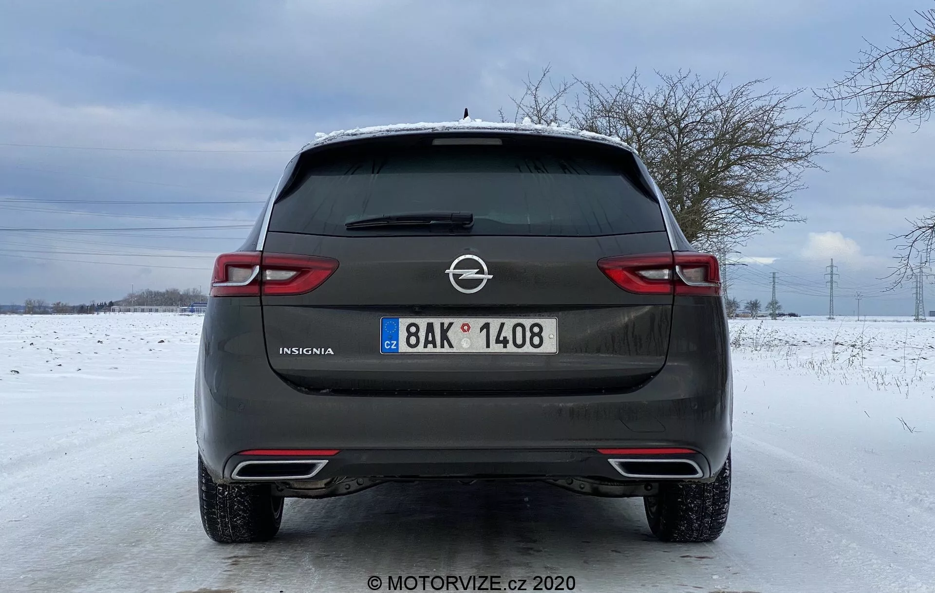 Bakre vy av en 2020 Opel Insignia Sports Tourer parkerad på en snötäckt väg. Den har en metallgrå finish, Opel-logotyp, modellnamnet 'Insignia', europeisk-stil registreringsskylt, bakre avfrostare med vindrutetorkare, dubbla avgasrör och en vinterbakgrund med ett snötäckt fält och nakna träd under en molnig himmel.