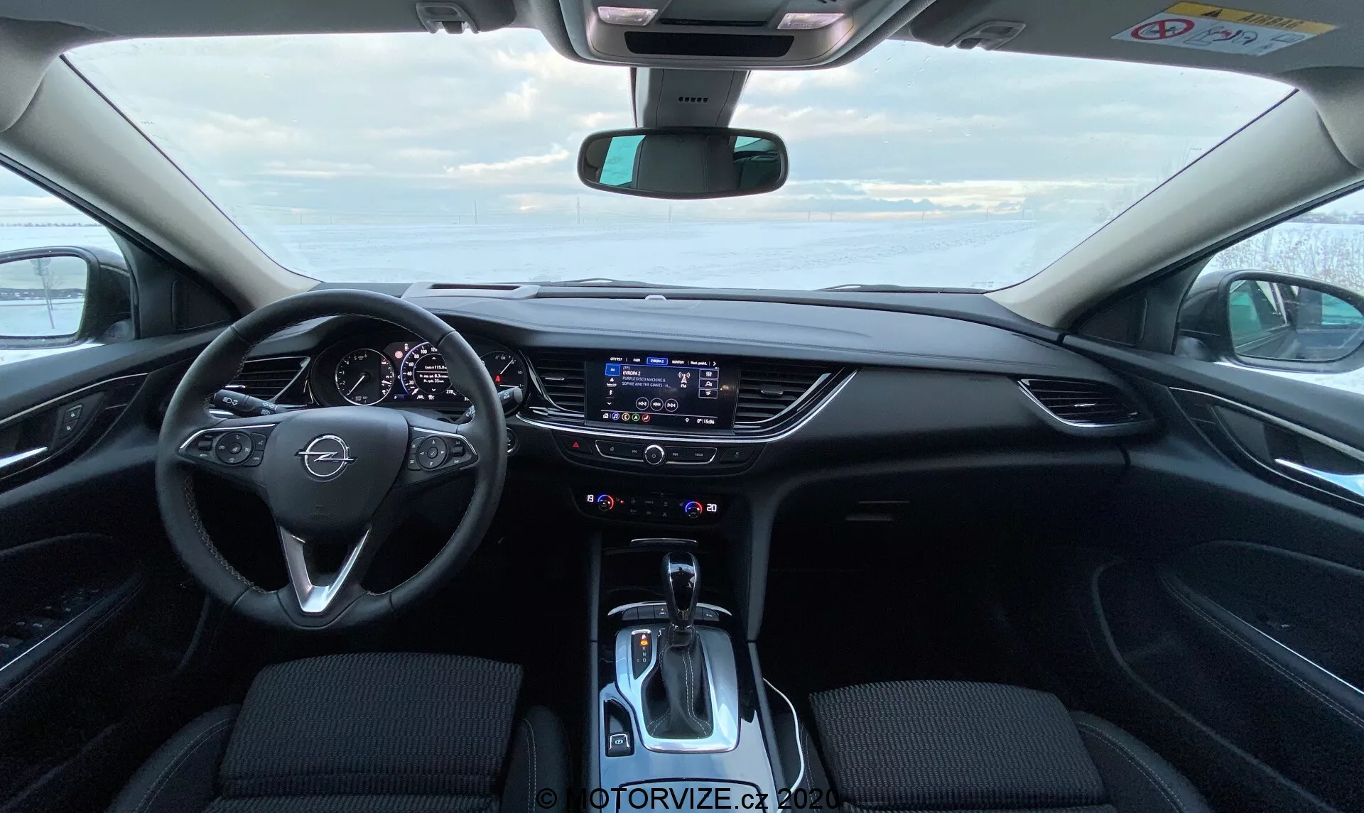 Interiörvy av en modern Opel Insignia Sports Tourer 2020 från förarsätets perspektiv, som visar ratten med integrerade multimediakontrollknappar, en delvis synlig modern instrumentpanel, ett stort flush-monterat pekskärmsystem för infotainment på instrumentbrädan, tvåzoners klimatkontroller, en modern växelspak och mörka tygklädsel i förgrunden.