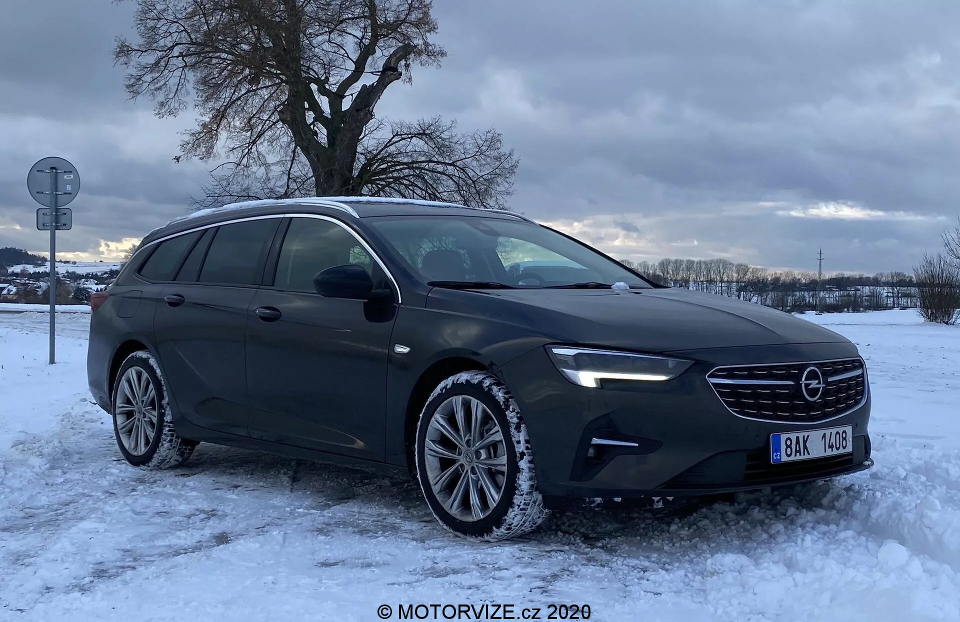 Bilden visar en Opel Insignia Sports Tourer 2020 fotograferad från en trekvarts framprofil. Vinkeln visar tydligt frontgrillen med märkesemblemet, eleganta strålkastare kanske med LED-teknik, motorhuvens konturer och förarsidan inklusive fram- och bakdörrar samt sidospeglar. Den kombimodellens förlängda form och förlängda taklinje framhäver dess större lastutrymme. Mot en vintrig bakgrund med snö på marken och mulen himmel, är bilen parkerad på en snötäckt yta med synliga hjulspår. Frontstötdämparen har dimljus och alufälgarna är utrustade med vinterdäck. Bilens mörka metalliska lack och reflekterande fönster tyder på privat- eller UV-behandling. En sidoblinker integrerad i sidospegelhuset förbättrar säkerheten genom att signalera sväng- och körfältsändringsavsikter.