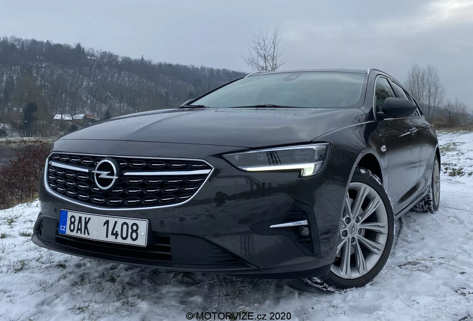 Bilden visar en svart Opel Insignia Sports Tourer 2020 fångad från en vinkel av tre kvarts front, vilket framhäver fordonets vänstra sida. Denna perspektiv inkluderar främre stödfångare, grill med framträdande logotyp, motorhuv, vänstra sidans strålkastare med avancerad LED-teknologi, en del av vindrutan, samt förarens främre hjul och dörr. Synliga är fler-ekrade lättmetallfälgar och sidospegel med integrerad blinkers. Fotograferad i en lätt nedåtvinkel i ett vinterlandskap med lätt snöfall eller frost på marken och molnigt väder med tillräckligt omgivningsljus som framhäver designen och möjliga aerodynamiska och kylfunktioner av främre stödfångarens luftintag.