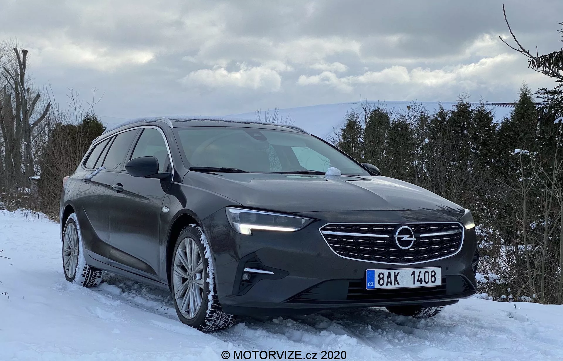 Opel Insignia 2020 Sports Tourer, en modern kombi, parkerad på ett snöigt underlag med vinterträd och en delvis molnig himmel i bakgrunden. Framifrån i trekvartsvinkel visas fordonets eleganta grill med tillverkarens logotyp, LED-strålkastare och aerodynamiska linjer på motorhuven. Flerekerfälgar med vinterdäck, lämpliga för snöförhållanden, är synliga. Bilens mörka färg, troligen grafit eller mörkgrå med en glansig finish, reflekterar naturligt ljus och framhäver en design fokuserad på praktiskhet och komfort snarare än sportiga attribut.