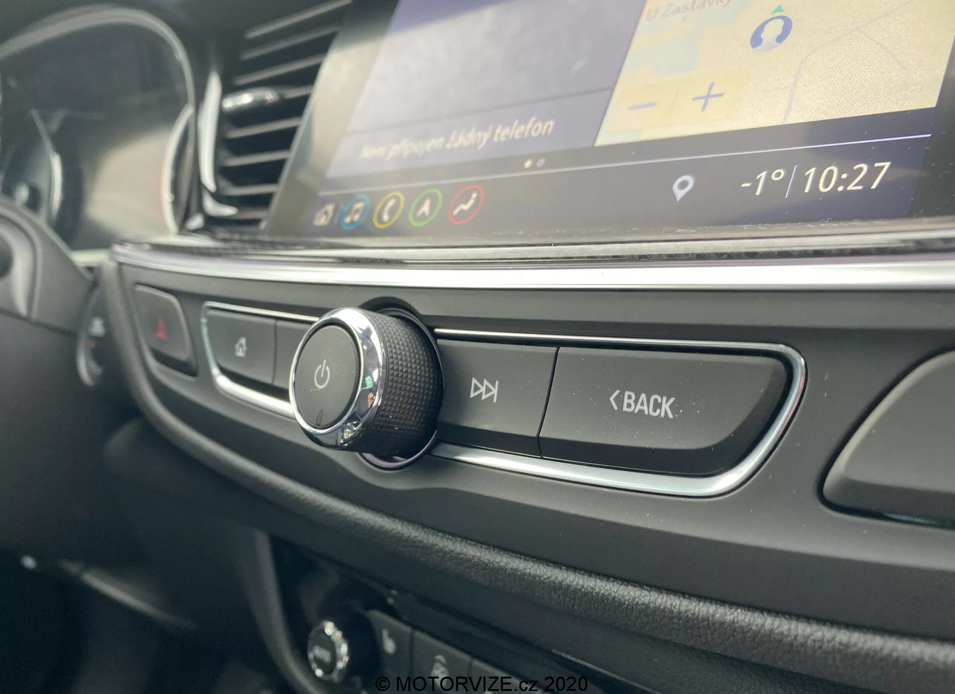 Närbild av interiören i Opel Insignia Sports Tourer 2020 från förarens sida, med fokus på den roterande kontrollratten, matt svarta knappar för ljudkontroll och en oskarp infotainment-skärm som visar ett grafiskt användargränssnitt med tid och temperatur.