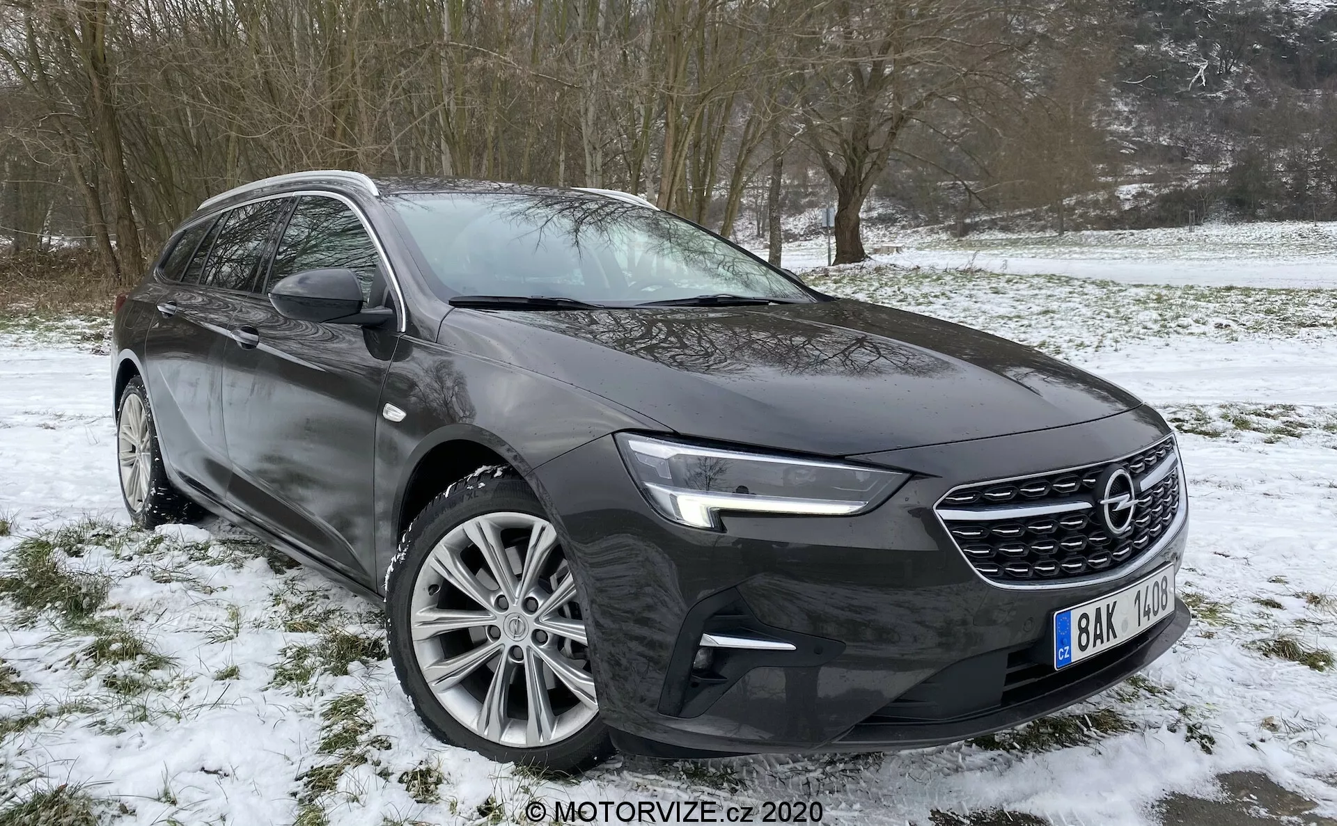 Bild av en modern personbil från 2020, en Opel Insignia Sports Tourer, parkerad på en snöig och gräsbevuxen yta, taget från trekvarts vinkel mot vänster framsida. Bilden framhäver de främre strålkastarna med LED-dagsljus, en framträdande grill med horisontellt mönster och en del av stötfångaren. Motorhuven har sportiga konturlinjer och den infällda sidobackspegeln har inbyggda riktmärken. Flerekrade aluminiumfälgar bidrar till det exklusiva utseendet. Förarsidans profil visar form av fönstren, dörrhandtag och dynamiska karaktärslinjer, med de nedre delarna av däcken och fälgarna täckta av snö.