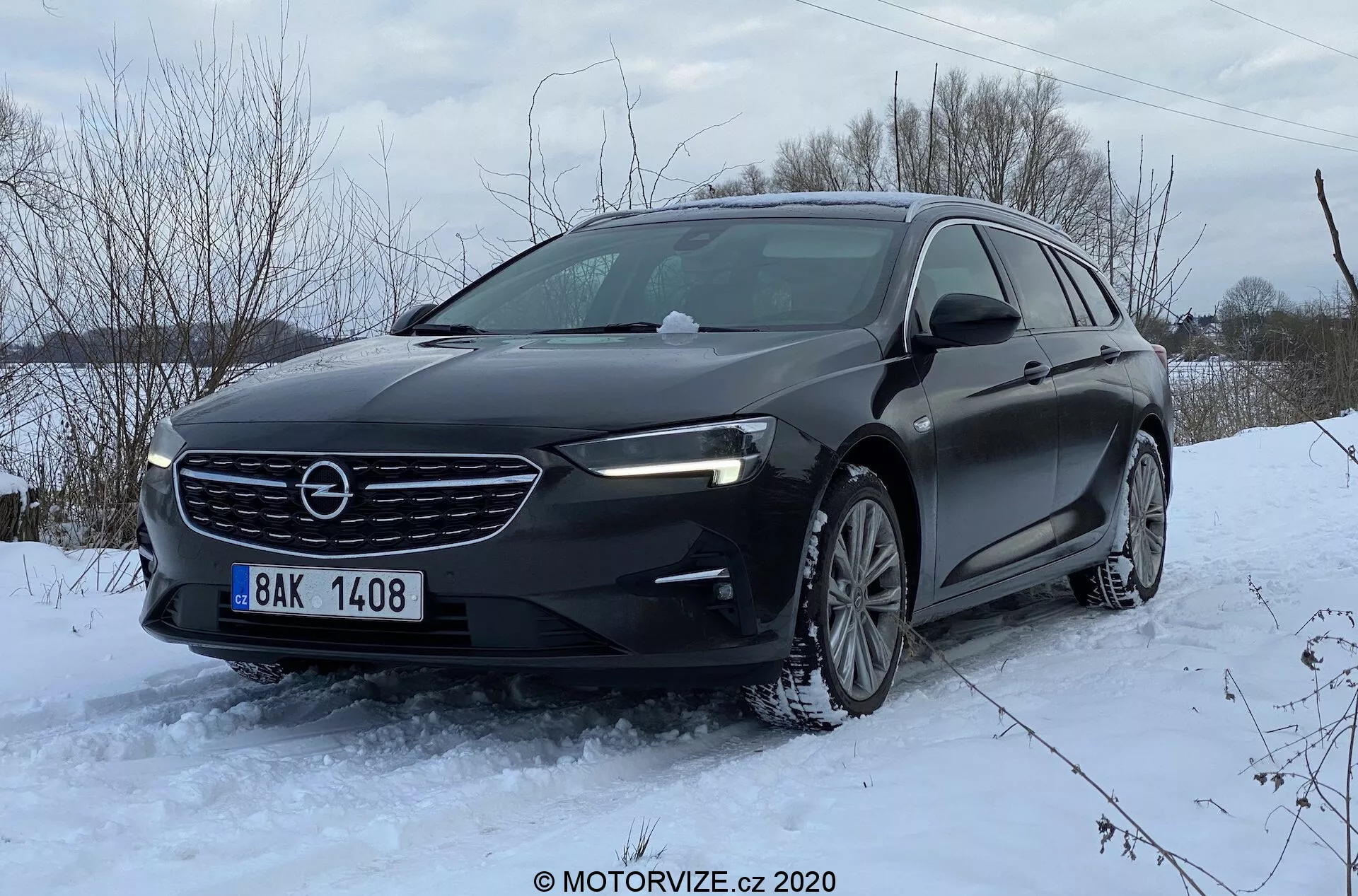Låg front trekvarts vinkel av en Opel Insignia Sports Tourer 2020, som visar den främre grillen med horisontella ribbor och centralt placerad logotyp, moderna eleganta strålkastare med LED-dagsljus, konturerad motorhuv, främre stötfångare med luftintag och troliga parkeringssensorer, lättmetallfälgar och vinterdäck. Bilen, som står stilla i ett snötäckt landskap, fotograferad i dagsljus, framhäver dess vinterkörningsegenskaper och dynamiska hållning.