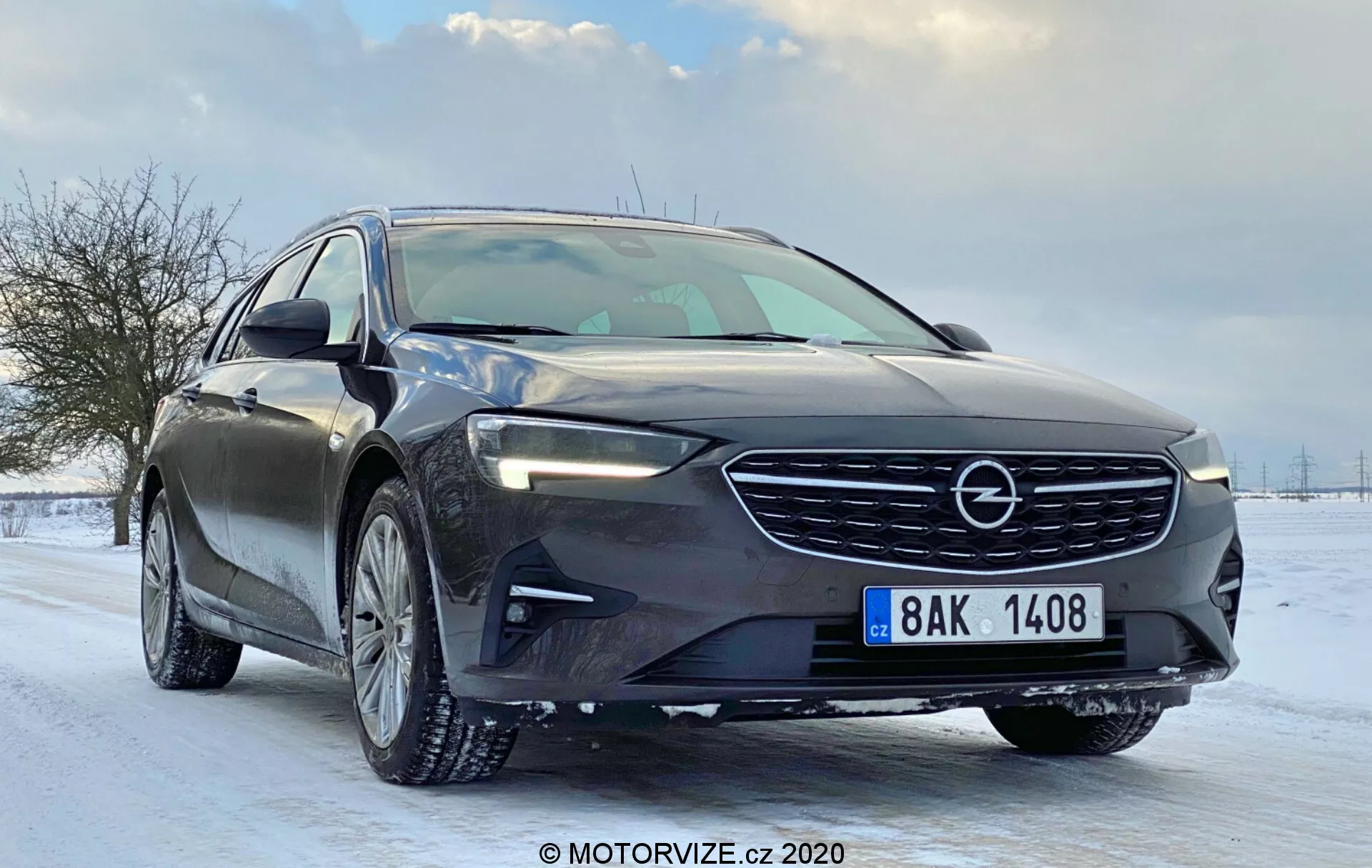 Bild på en modern bil (Opel Insignia 2020 Sports Tourer) från en låg frontal tredjedelsvinkel som visar dess framträdande frontgaller med märkets emblem, distinkt horisontellt mönster, LED-strålkastare med dagkörljus, elegant motorhuv med aerodynamiska konturer, lättmetallfälgar med vinterdäck, strömlinjeformade sidobackspeglar med integrerade blinkers, kromfönsterlister, synlig karaktärslinje genom dörrhandtagen, lutande vindruta, glansig mörk färg, snötäckt vägighetsmiljö, synlig främre registreringsskylt, vilket antyder avancerade tekniska egenskaper.