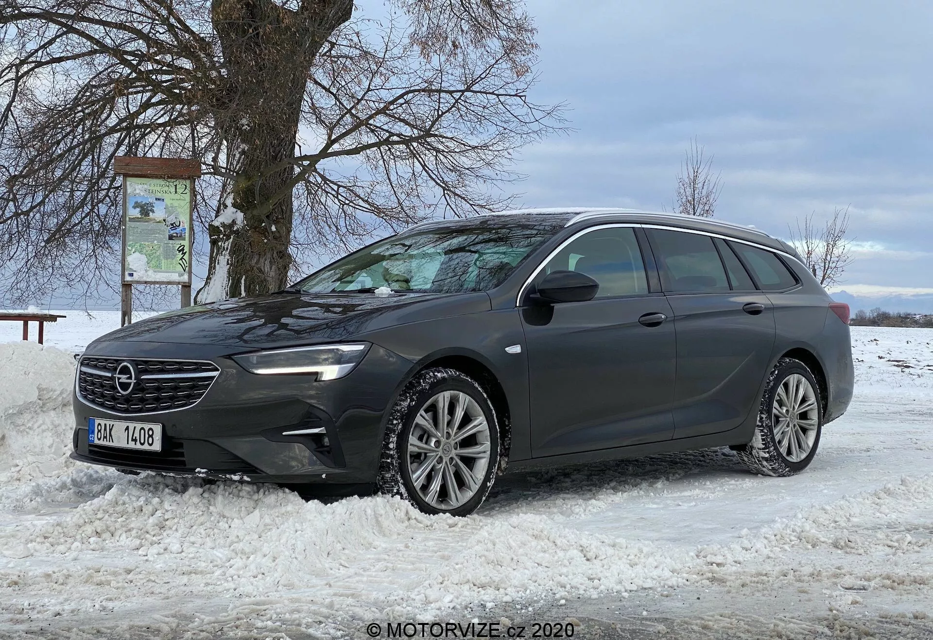 Denna bild visar en modern kombi, Opel Insignia Sports Tourer 2020, parkerad på ett snötäckt underlag. Bilen är placerad diagonalt och ger en tydlig vy av sin främre profil och hela förarsidan. Vinkeln på bilden är en trekvart främre vy, som framhäver grilldesignen, de eleganta LED-strålkastarna, den främre stötfångaren och de flerfälgade aluminiumhjulen. Bilens längd, fönsterkonfiguration och takrälsystem är synliga och antyder lastnytta. En sidospegel, troligen utrustad med avancerad teknik, och en naturskön bakgrund med ett lövlöst träd och en molnig himmel förbättrar scenen.
