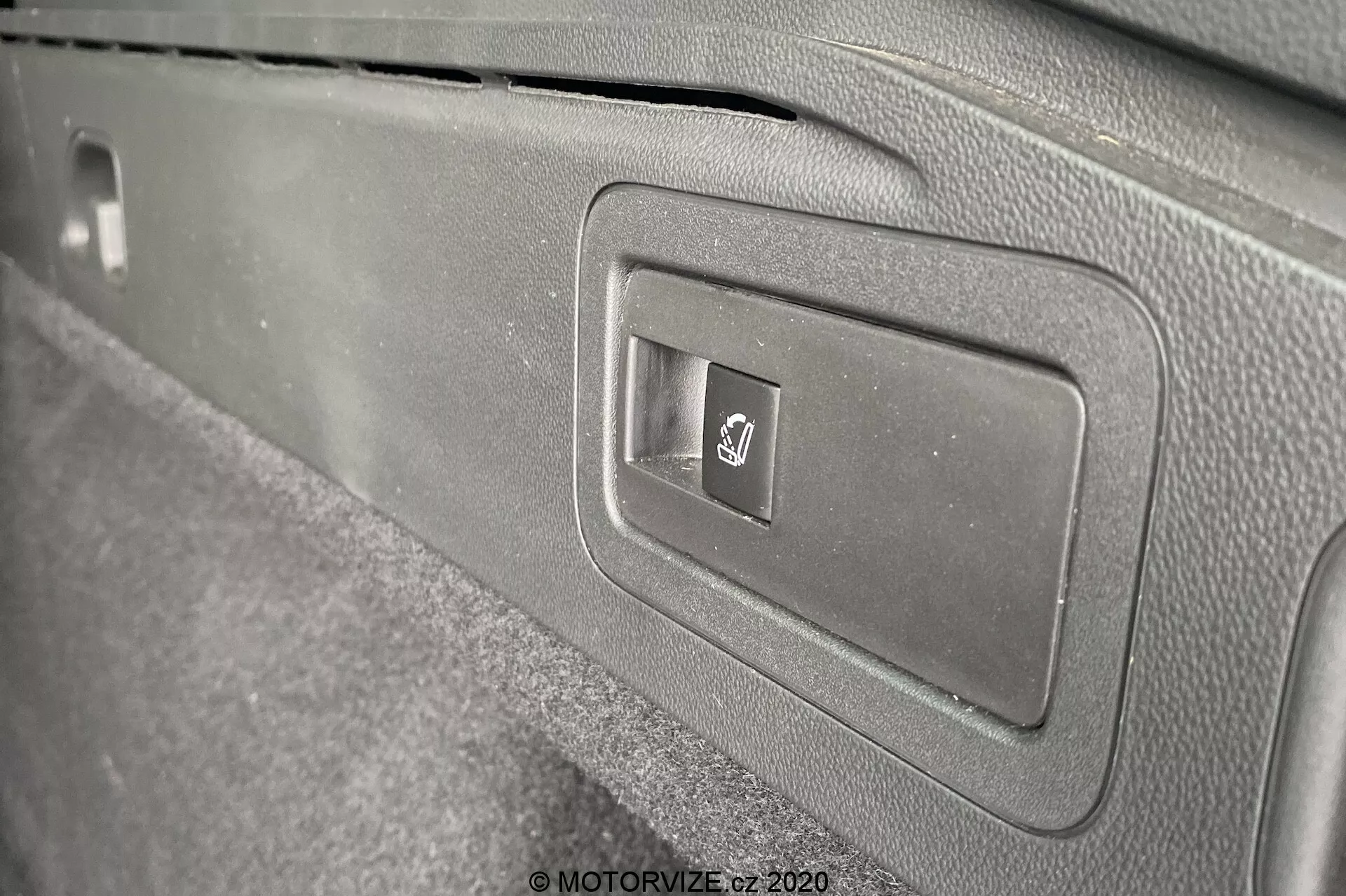 Närbild på inre panelen i 2020 Opel Insignia Sports Tourer, som fokuserar på en rektangulär plastlucka med en elektronisk bagageluckeöppningsknapp. Knappen är märkt med den universella bagageluckesymbolen. Bilden, tagen i sned vinkel, visar panelens rena design med olika nyanser av grått samt en mattliknande textur runt knappen.