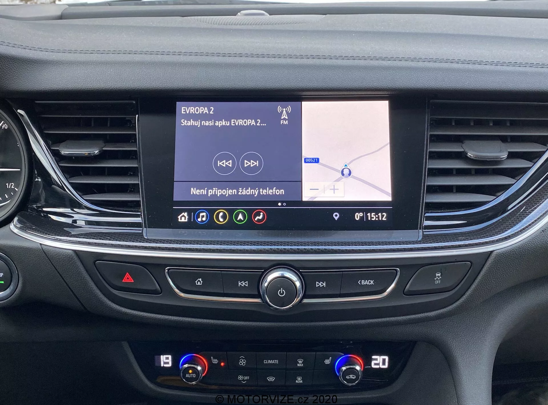 Vy över Opel Insignia 2020:s mittkonsol från förar- eller passagerarsätet, som visar infotainmentsystemet och klimatkontrollpanelen. Den färgglada LCD-pekskärmen, som är cirka 7 till 10 tum, visar en navigationskarta med slavisk text och etiketten 'EVROPA2', vilket antyder en position i Öst- eller Centraleuropa. Gränssnittet visar att ingen telefon är ansluten och tiden är 15:12. Nedanför finns horisontella luftventiler och knappar för multimediekontroll, varningsblinkers och andra inställningar, följt av ett dubbelzonigt klimatsystem som visar temperaturerna 19 och 20 grader Celsius. Den eleganta, moderna designen betonar den avancerade biltekniken för en bekväm användarupplevelse.