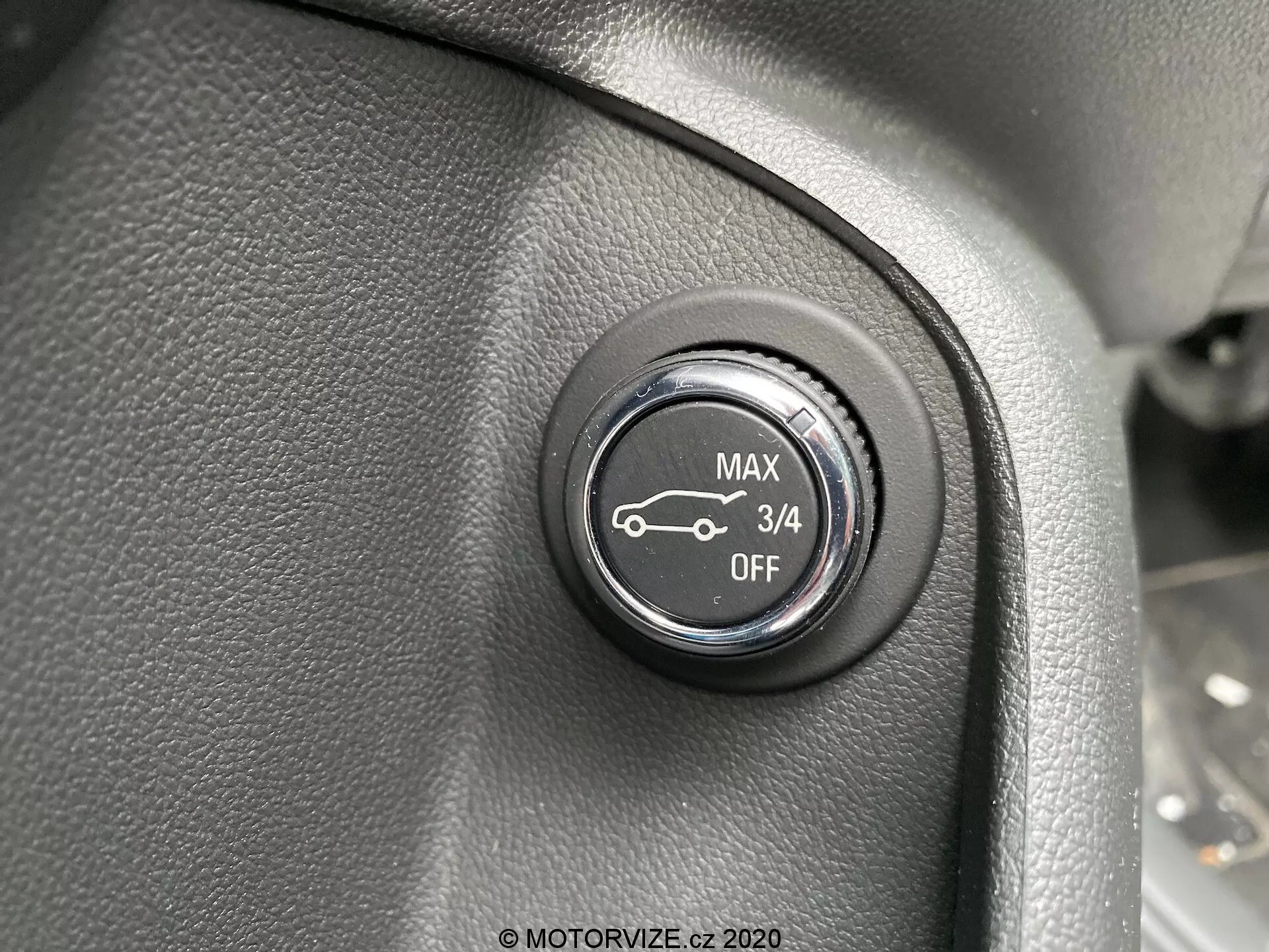 Närbild av en kontrollratt i Opel Insignia 2020:s interiör. Placerad på en texturerad yta, sannolikt en del av instrumentpanelen eller sidopanelen nära ratten, är den svarta ratten med vita markeringar som visar fyra positioner: 'MAX,' en bilsymbol med en uppåtpil, '3/4' och 'OFF.' Detta antyder att den justerar en funktion som strålkastare eller fjädringsinställningar. Fotot fokuserar på rattens framsida, något uppifrån, med ett grunt skärpedjup och jämn belysning.