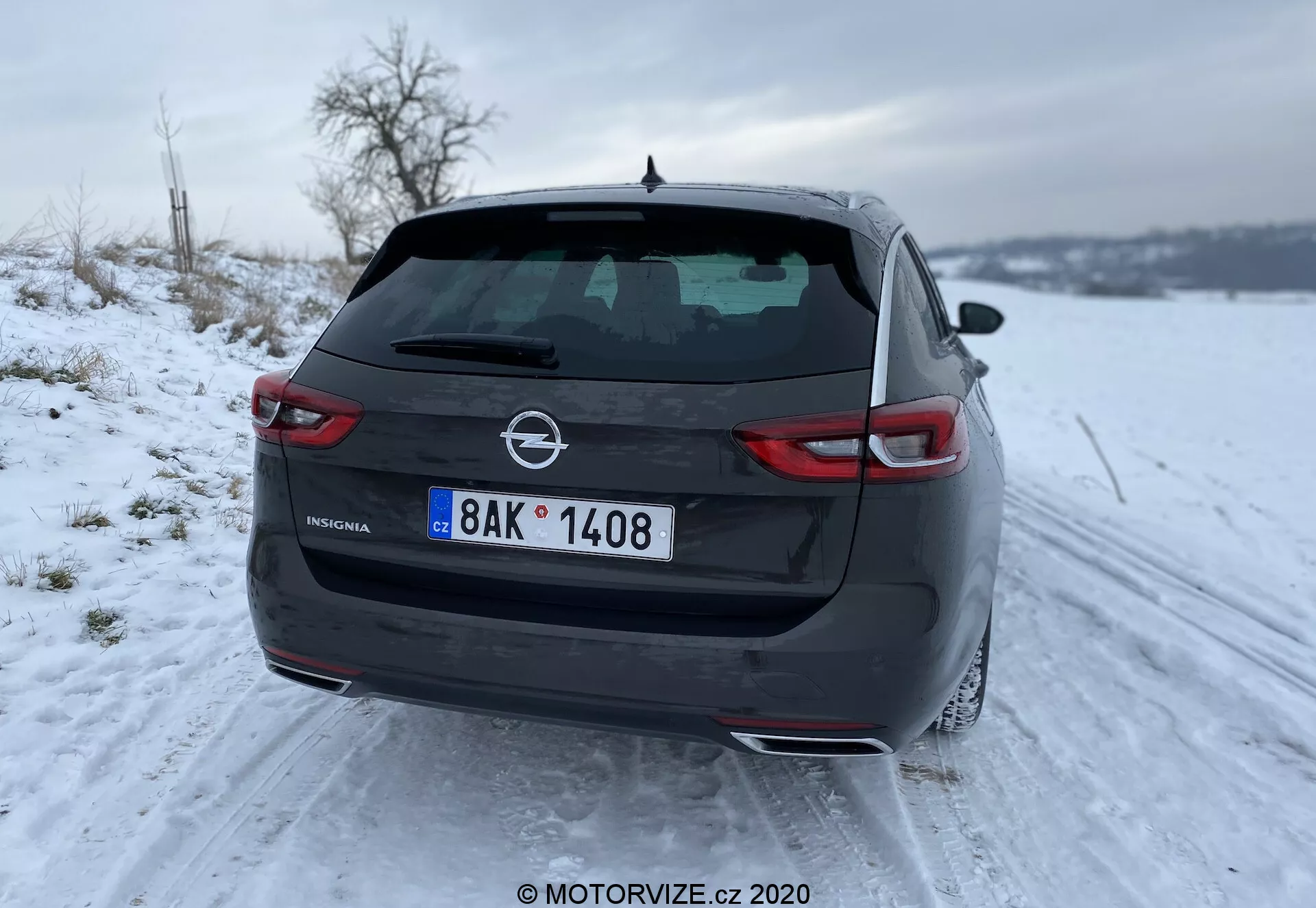 Bakåtsikt av den eleganta och moderna Opel Insignia 2020 Sports Tourer i ett snötäckt landskap, som visar smala LED-bakljus, dubbla avgasrör, bakruta med avfrostningsfunktion, tredje bromsljus integrerat i en spoiler och shark-fin antenn på taket. Bilden är tagen från en låg vinkel med fokus på den vänstra sidan, betonar insignia och tillverkarens märke på bagageluckan.