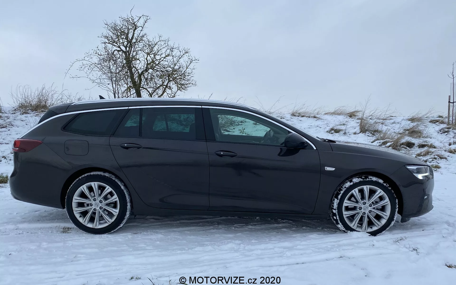 En sidoprofil av en mörkfärgad 2020 Opel Insignia Sports Tourer kombi, parkerad på en snötäckt yta. Fotot, taget under en mulen dag med dämpat naturligt ljus, visar fordonets moderna designelement, inklusive flerspåkiga aluminiumfälgar, lågprofildäck och eleganta tonade fönster. De integrerade körriktningsindikatorerna i sidospeglarna, de spolande dörrhandtagen i matchande karossfärg och en takspoiler med möjliga aerodynamiska fördelar är också synliga. Bilen saknar synliga emblem eller märken, och inga externa kameror eller sensorer går att urskilja från denna vinkel. De mulna förhållandena dämpar något designens detaljer, men bilens urban-familjeorienterade stil förblir tydlig.