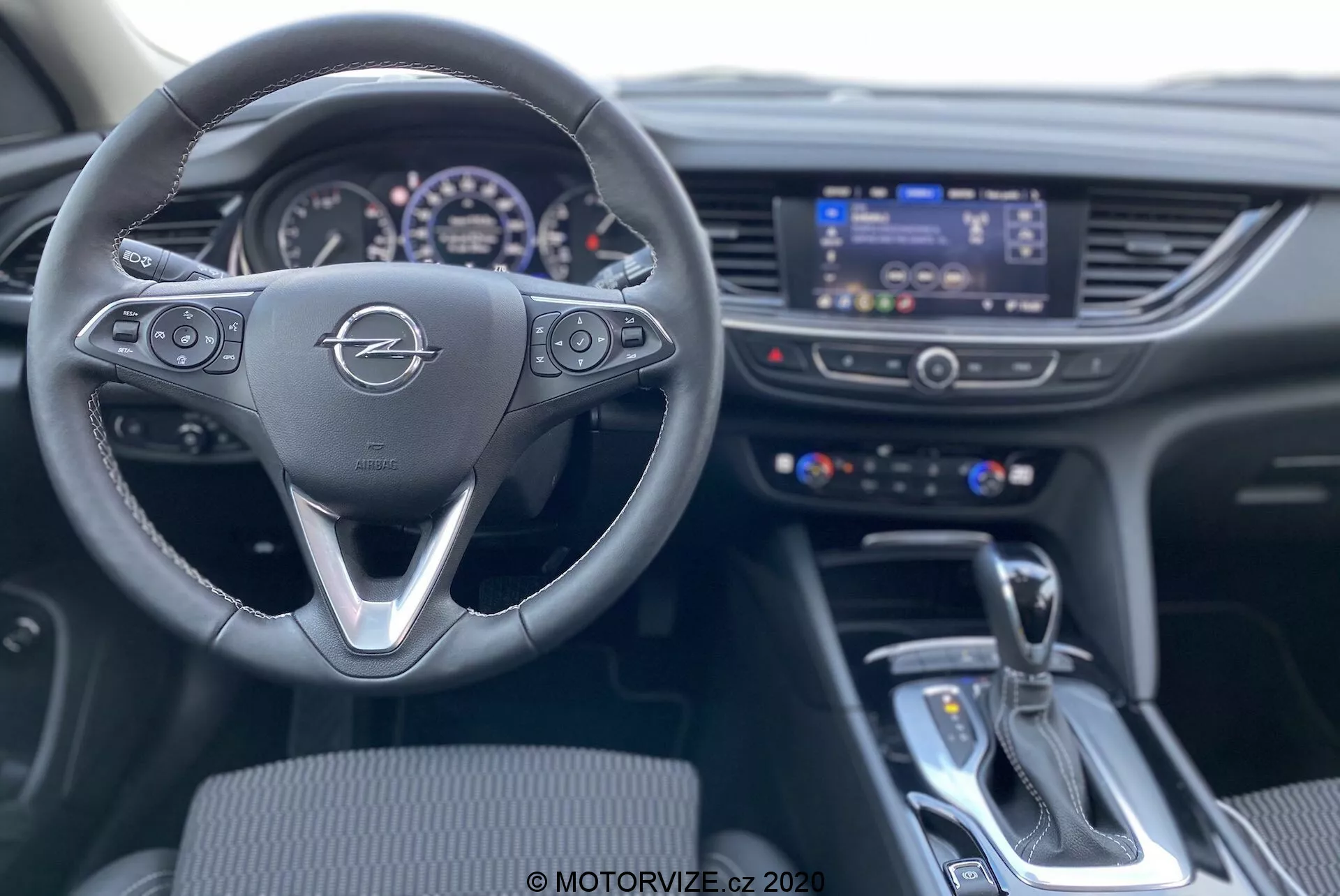 Inre vy från förarsätet i en Opel Insignia Sports Tourer 2020, med fokus på den treekrade ratten med multifunktionskontroller, det upplysta instrumentbrädet och den centrala infotainmentskärmen omgiven av klimatkontrollomkopplare. Bilden visar även växelspaken för automatväxellådan och närliggande kontrollenhet på mittkonsolen. Interiören är modern och framhäver funktionalitet och teknologisk integration.