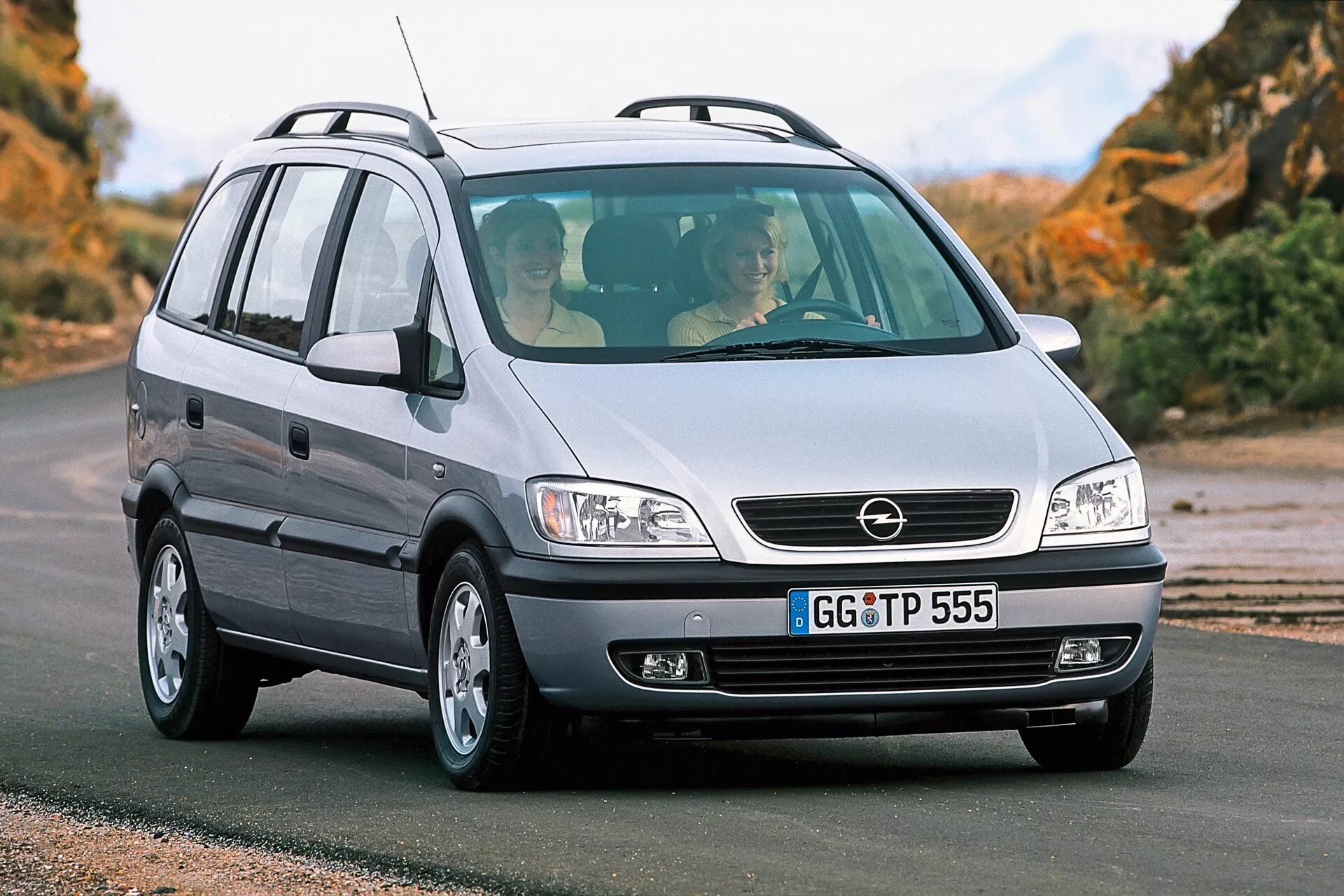 Fotografi av en Opel Zafira från 1999, en kompakt MPV, från en låg främre tredjedelsperspektiv. Bilden framhäver bilens grill med ett centralt emblem, stora strålkastare som troligtvis använder halogenljuskällor, slät motorhuv, främre stötfångare med dimljus, lättmetallfälgar och passagerarsidan på fordonet med en antenn på bakre delen av taket. Fordonet är på en asfalterad väg med ett naturligt landskap i bakgrunden. Genom den främre vindrutan kan man se två passagerare, vilket reflekterar fordonets interiördesign och rymlighet.