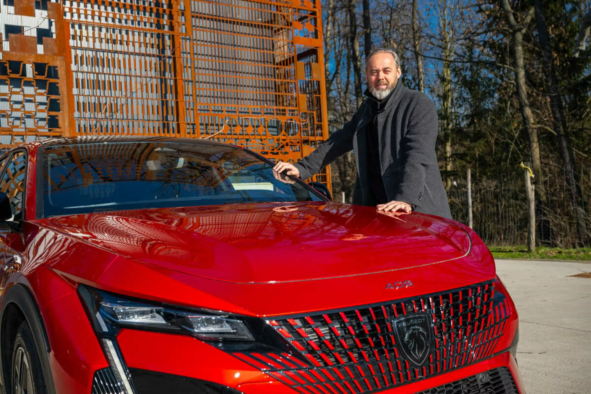 Röd modell av Peugeot 408 från 2023 som visar sin främre vänstra kvart i dagsljus, framhävande av detaljerade LED-strålkastare, geometrisk grill och sportig, aerodynamisk huva med en man i en mörk kappa bredvid som placerar sin hand på huven