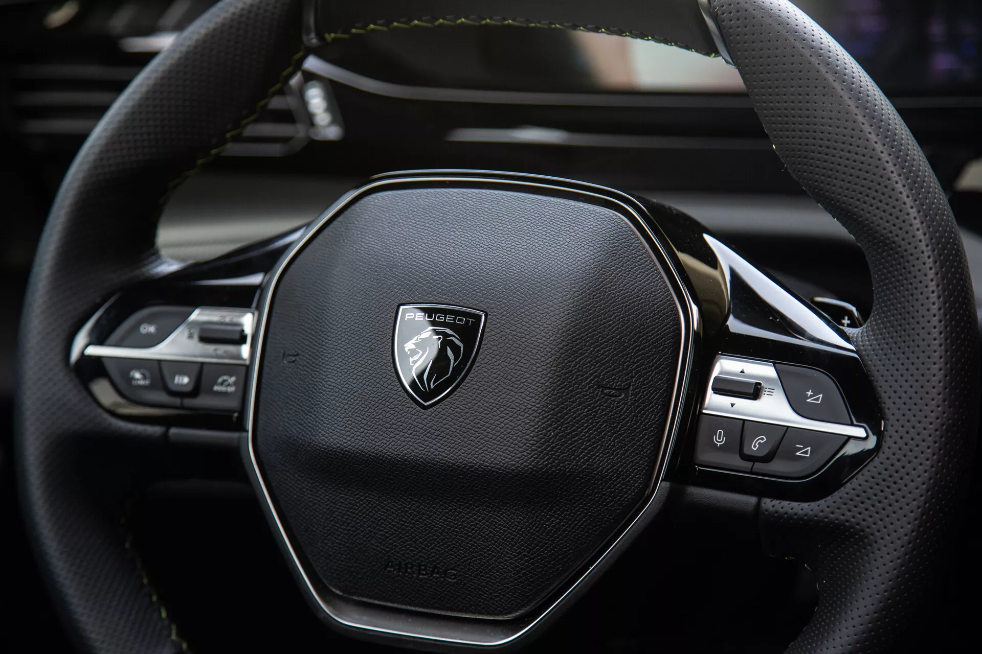 Närbild av Peugeot 408 2023 ratten som visar ergonomisk sexkantig design, lädersömmar, integrerade teknikreglage för ljud och kryssningsinställningar, med emblem på borstad metall.