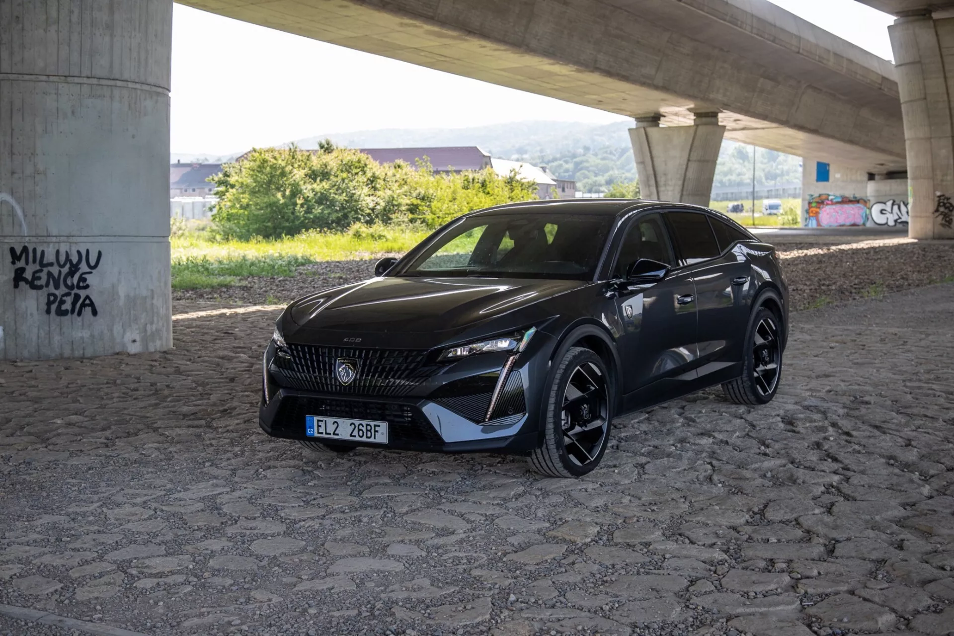 Peugeot 408 2023 modell fångad från en trekvarts vinkel som belyser dess släta LED-strålkastare, mönstrade grill och sportiga aerodynamiska frontstötfångare under naturligt dagsljus på ett asfalterat område under en viadukt