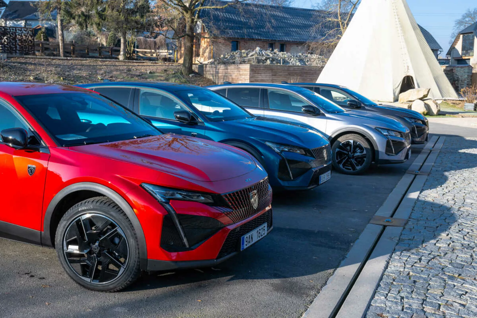 2023 års Peugeot 408-uppställning med röd bil i förgrunden med sportiga flerspakiga hjul och LED-strålkastare som antyder elektriska eller hybridmodeller, parkerad på asfalterat område intill kullersten