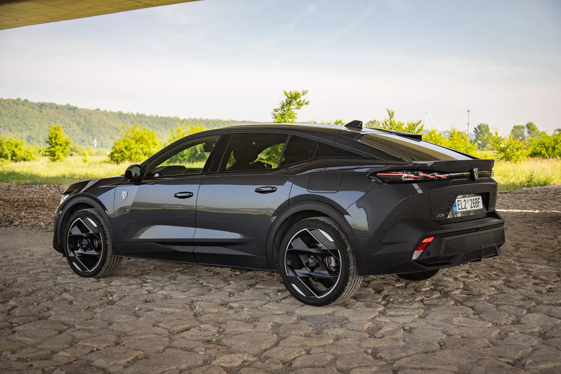 Crossover SUV Peugeot 408 från 2023 i mörk finish, fångar en tre fjärdedels bakre vy som framhäver aerodynamisk design med skarpa vinklar, sportig sluttande taklinje, stora svarta alufälgar, distinkta LED-bakljus och en diskret bakre spoiler på en kullerstensbelagd yta