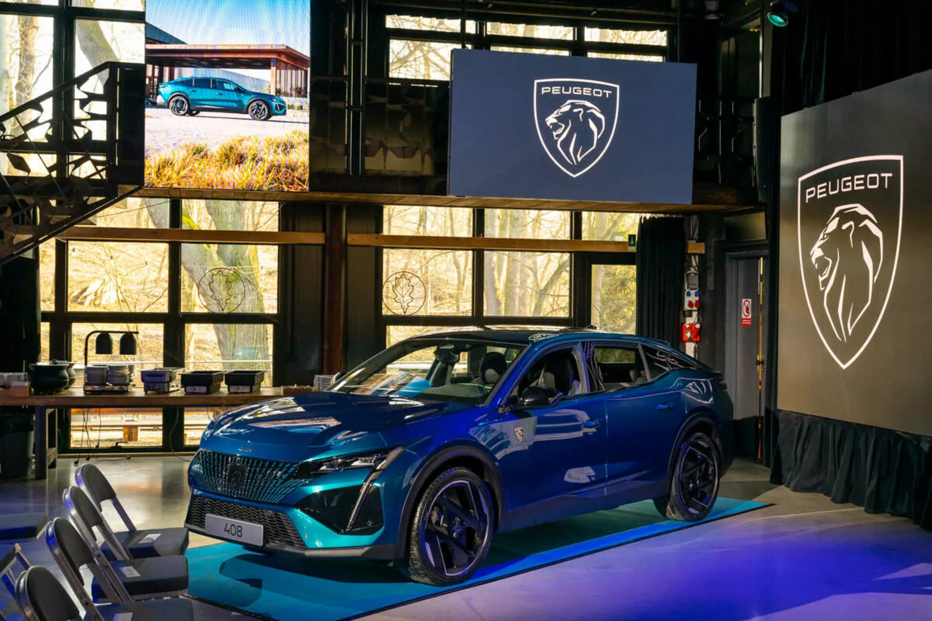 Peugeot 408 i blått från 2023 som visas på ett inomhusevenemang, med en distinkt grill, skarpa LED-strålkastare, sportigt konturerad motorhuv med luftintag, stora legeringshjul och en dynamisk sidoprofil med integrerade blinkers i dörrmonterade sidospeglar